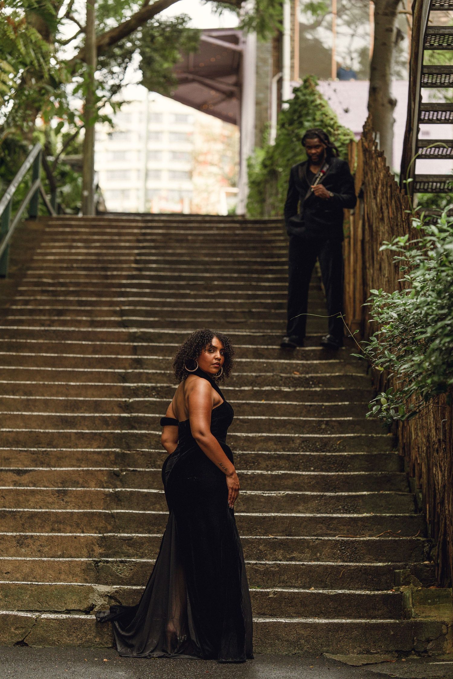 Woman in black gown on stone stairs with man standing in background.