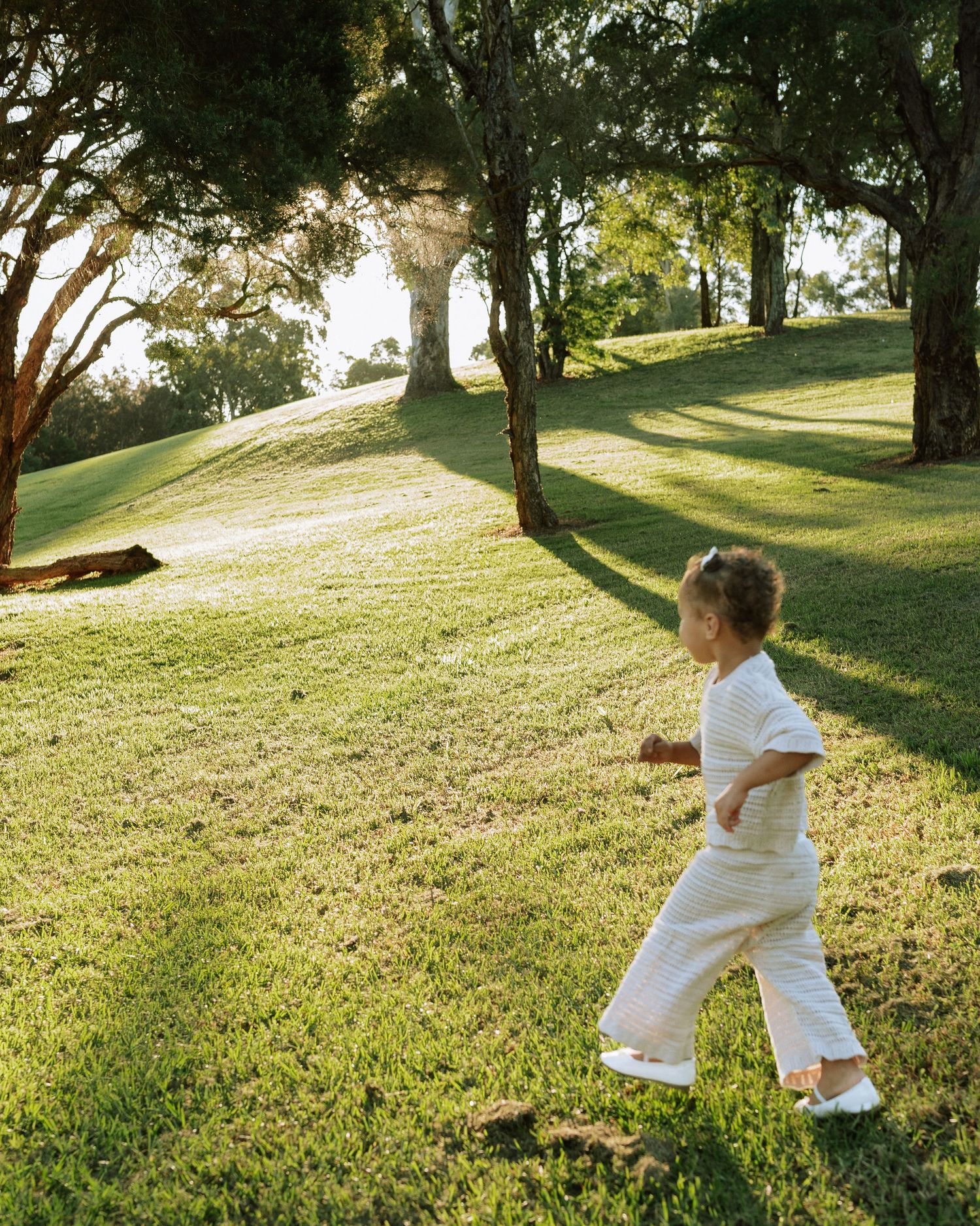 A young toddler in white outfit walks barefoot across lush green grass between tall trees.