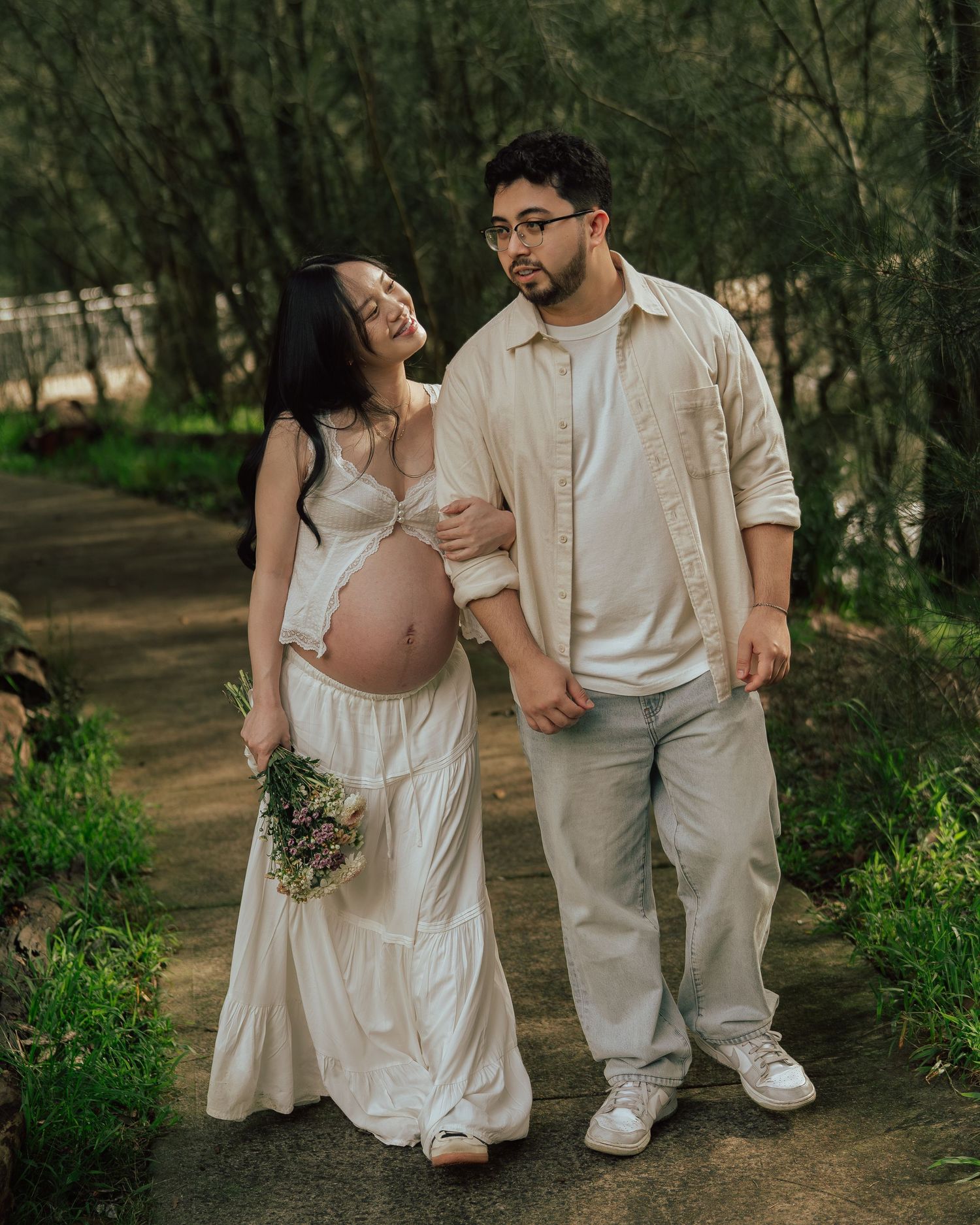 Expecting couple walks together on a lush green forest path, woman holding a wildflower bouquet.