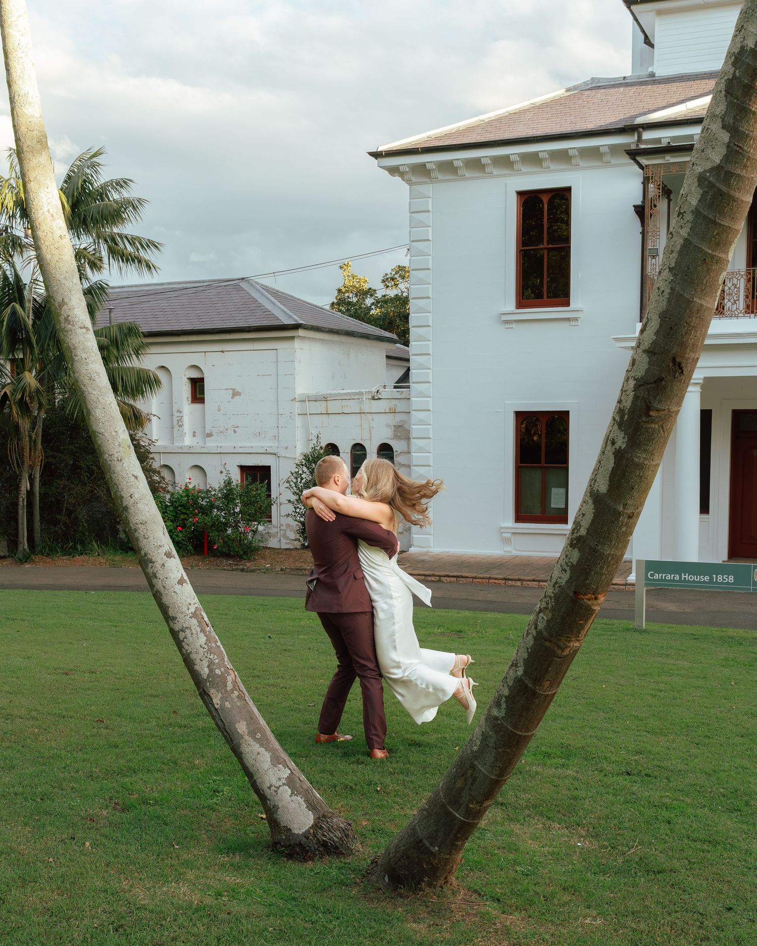 Groom lifts bride in white dress between leaning palm trees outside historic colonial building.