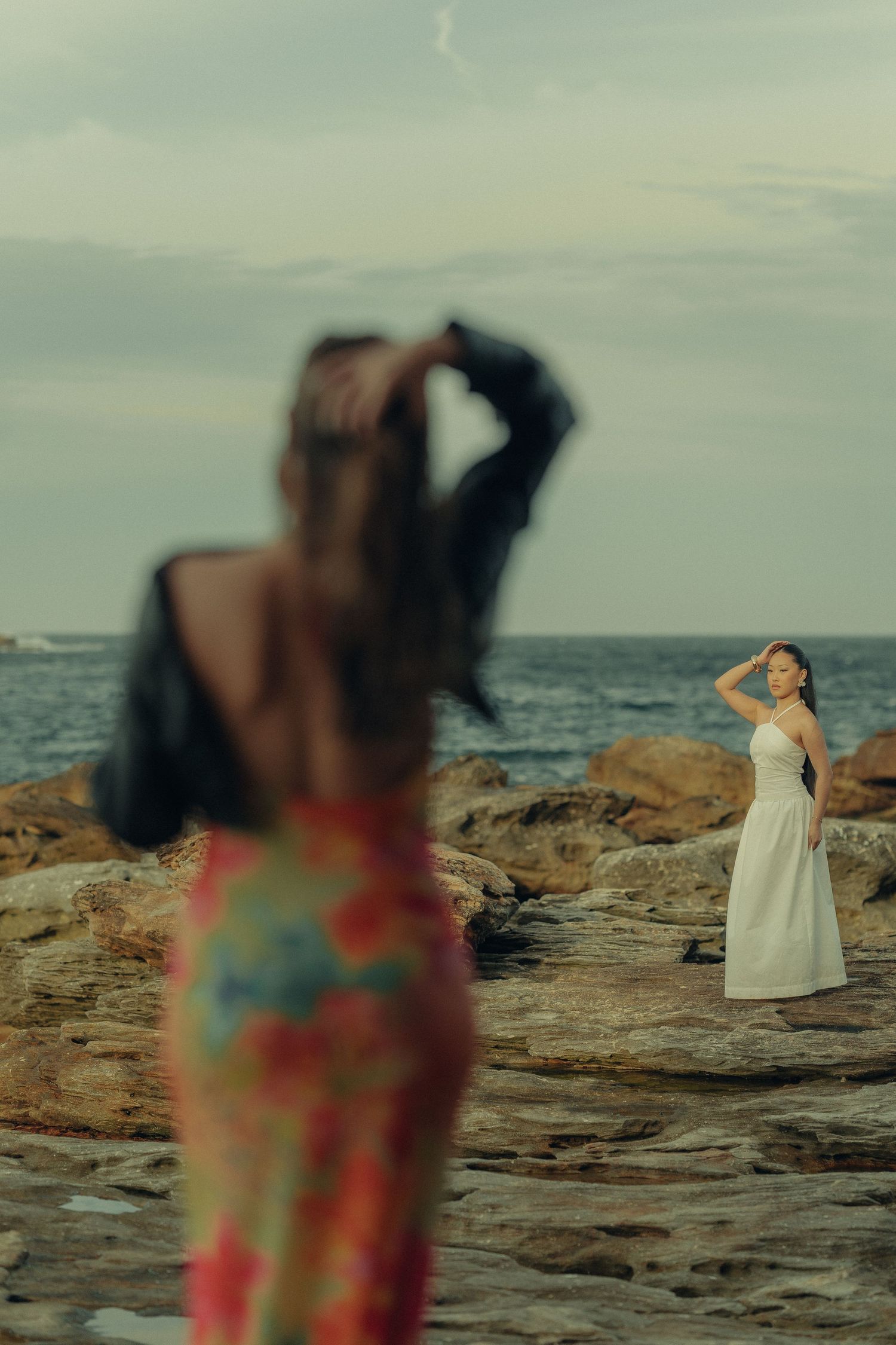 Two women in dresses on rocky shore, one in focus in white dress in background.