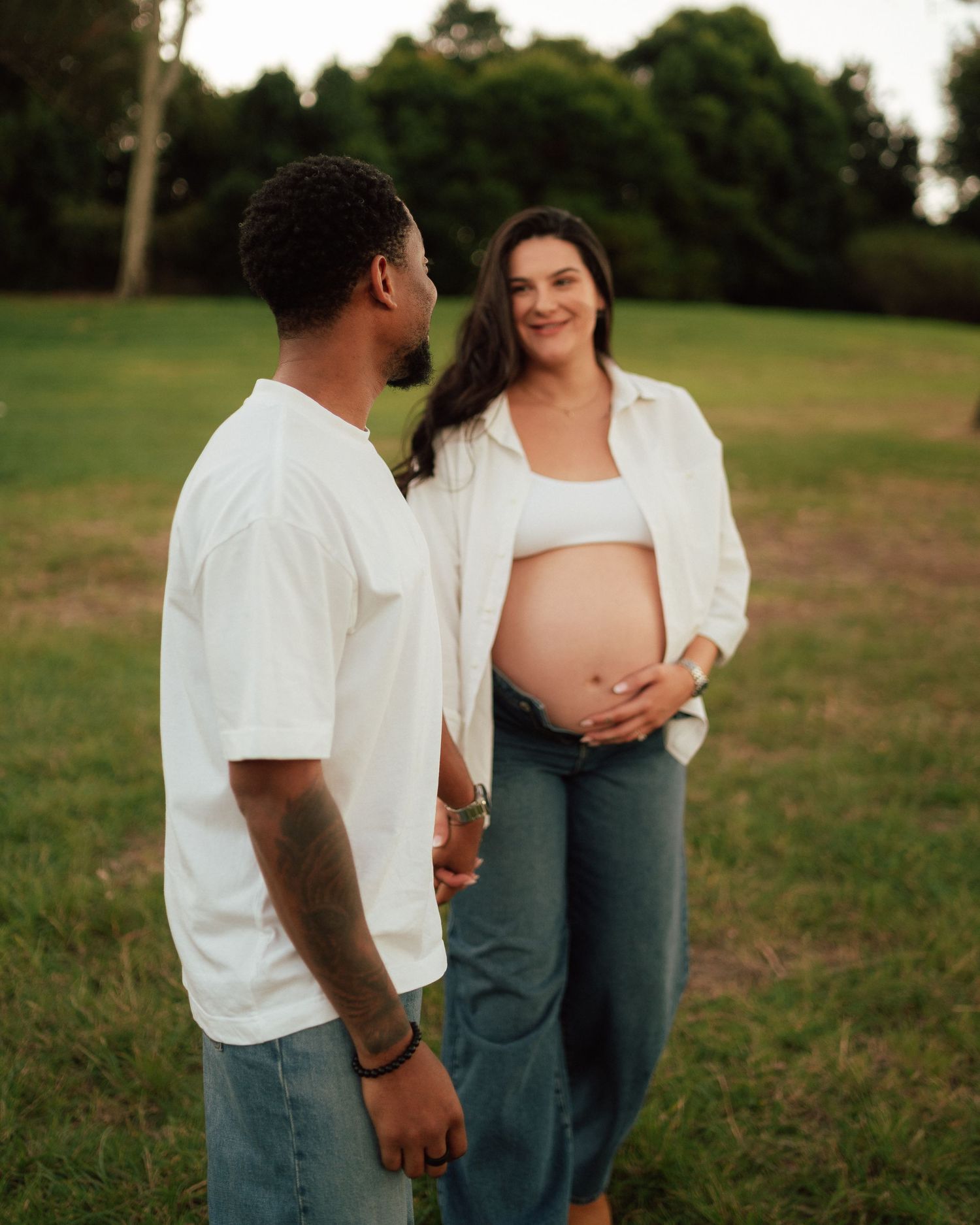 Couple facing each other smiling in a park at dusk, pregnant woman holding her bare baby bump outdoors.