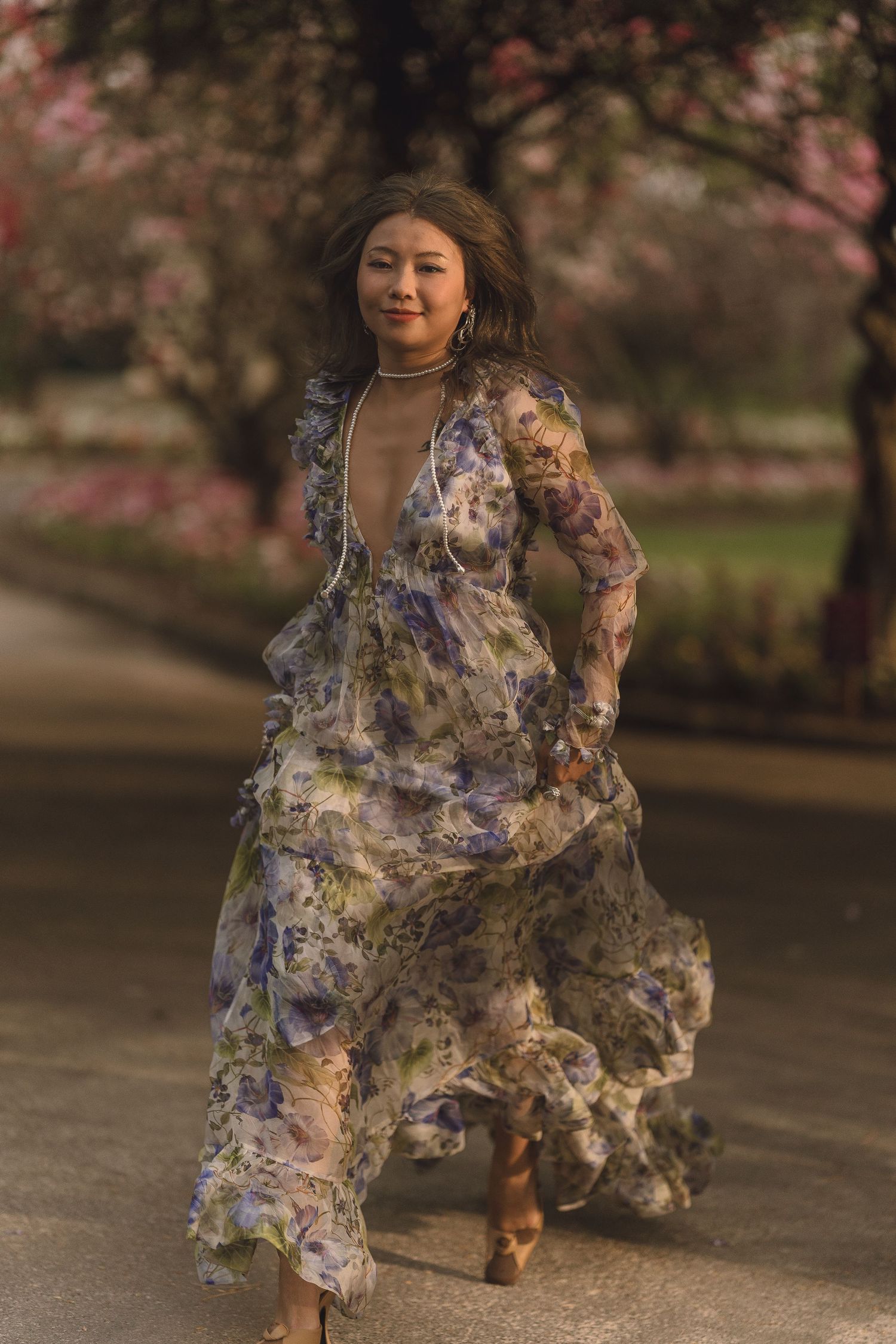 Woman in flowing floral lavender maxi dress walking among pink cherry blossom trees outdoors.