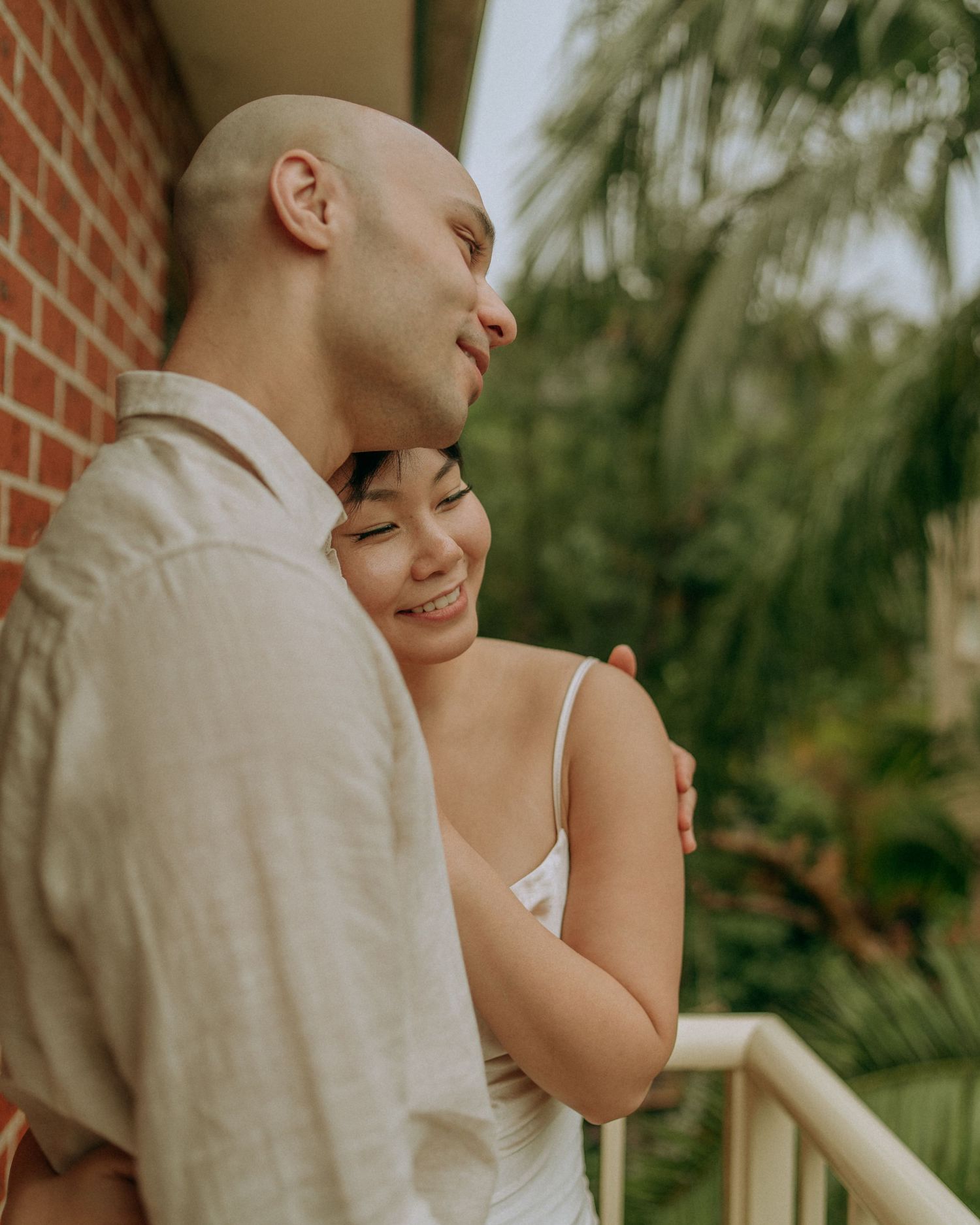 Couple sharing tender embrace on balcony with tropical palm trees in background.
