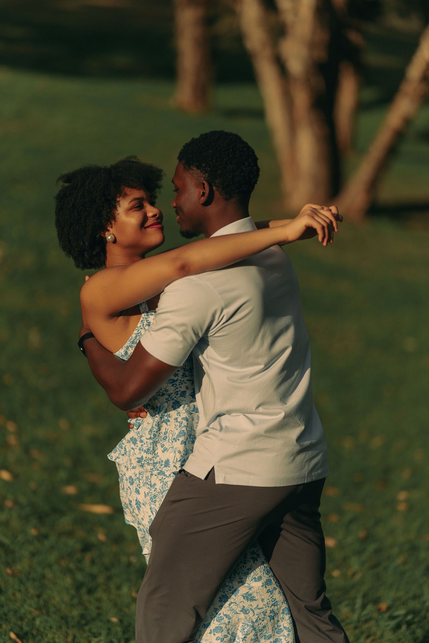 A couple dances together on a lush green lawn at golden hour, the woman smiling in a floral dress.