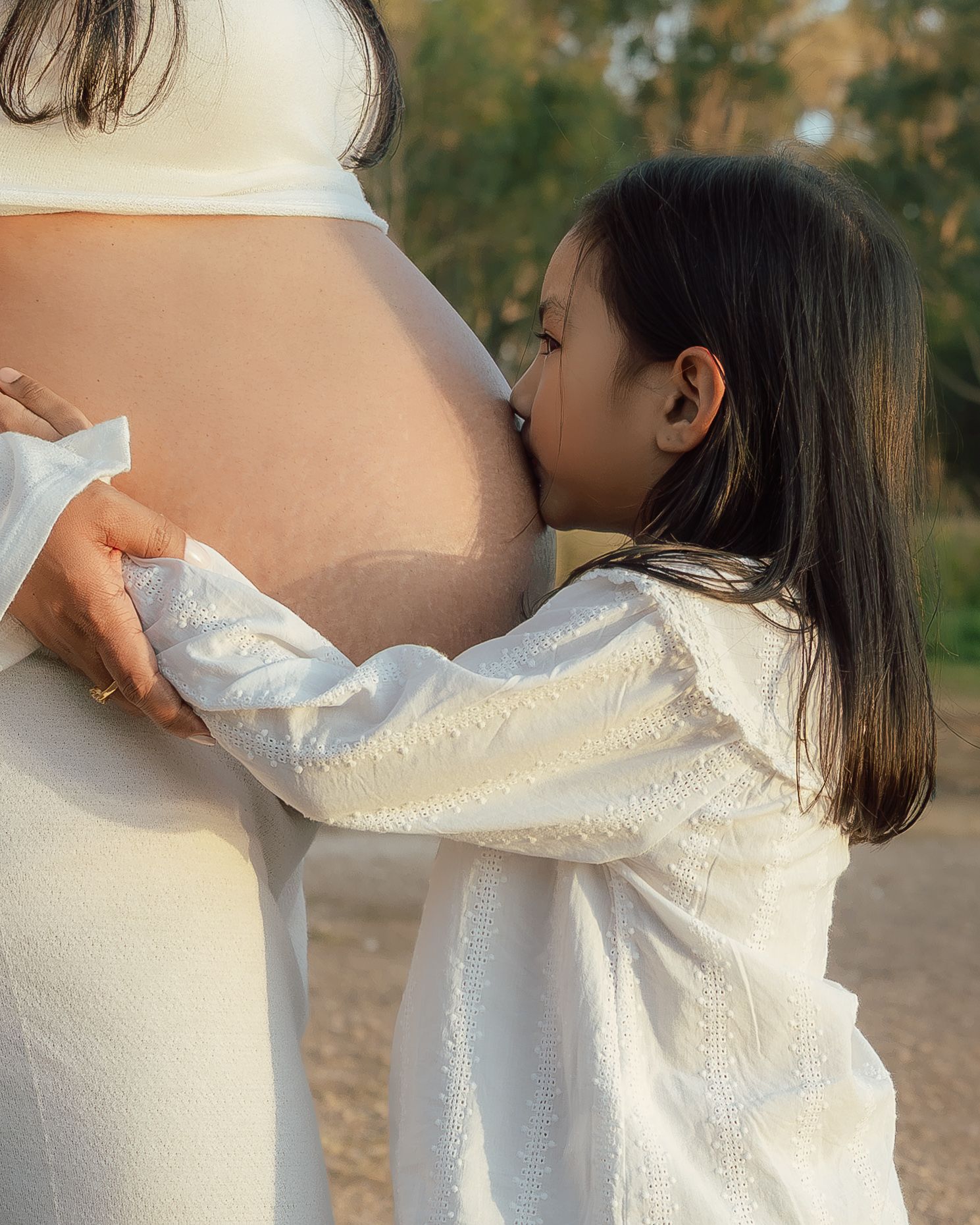 A young girl in white dress kisses her mother's pregnant belly outdoors in warm golden light.