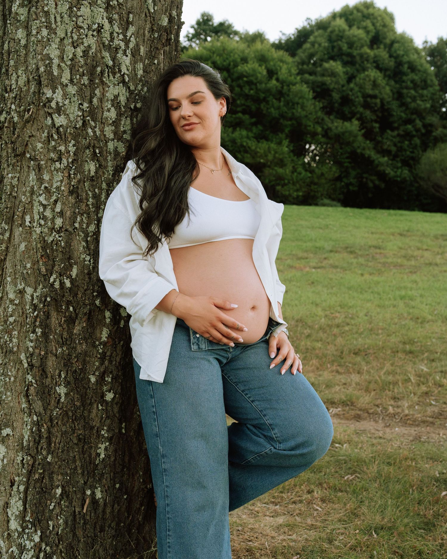 Beautiful pregnant woman in white shirt and jeans leaning against tree, cradling her baby bump outdoors.