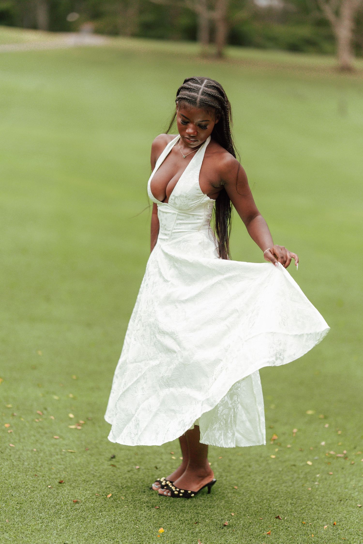 A young Black woman in a white halter dress twirls on a green lawn, lifting her skirt elegantly.