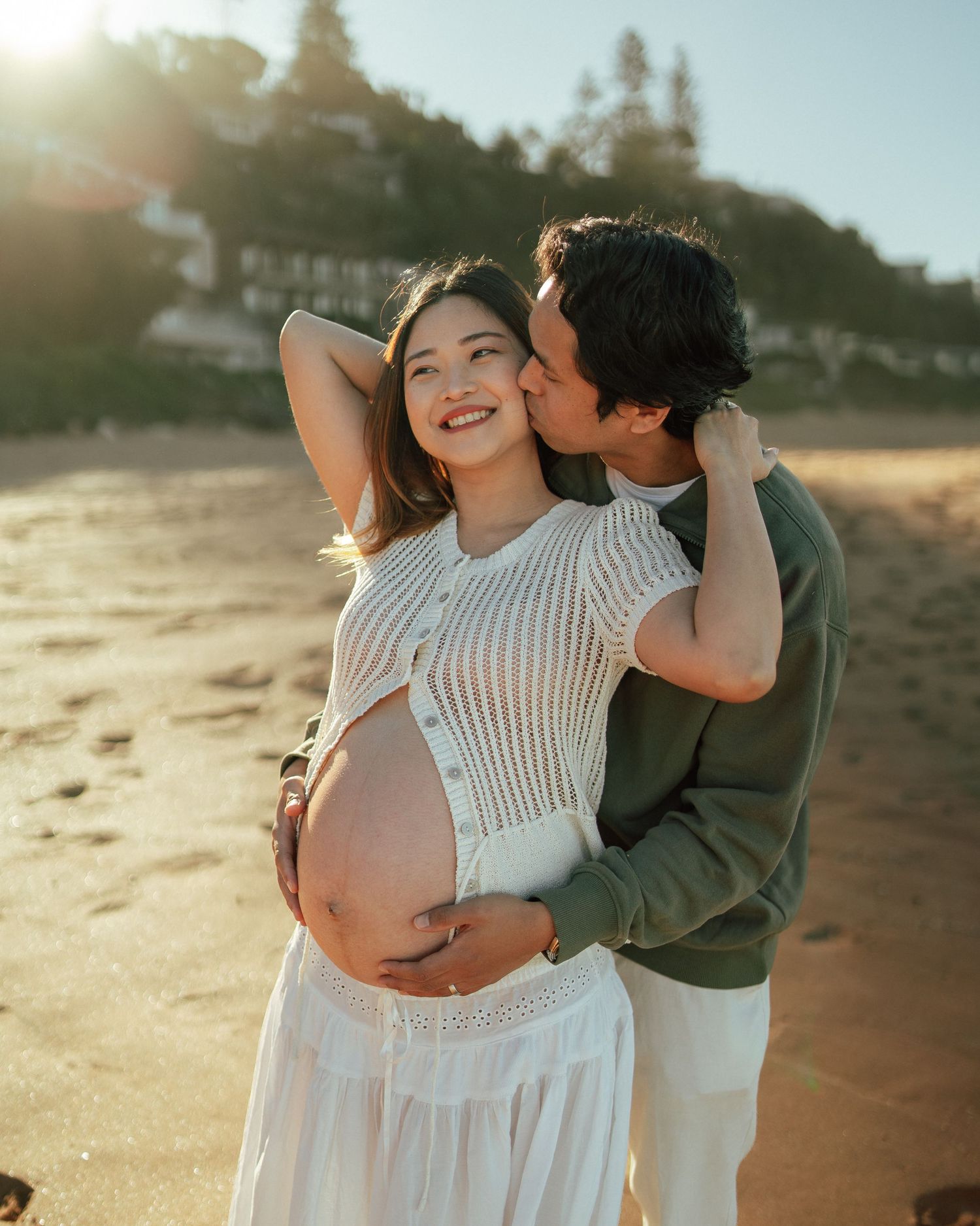 Happy pregnant couple embraces on a sunlit beach, partner kissing her cheek while holding her belly.
