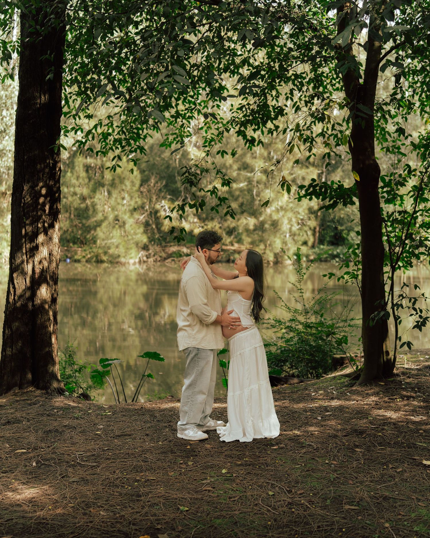 Couple sharing a romantic kiss near a tranquil forest lake during a maternity photo session.