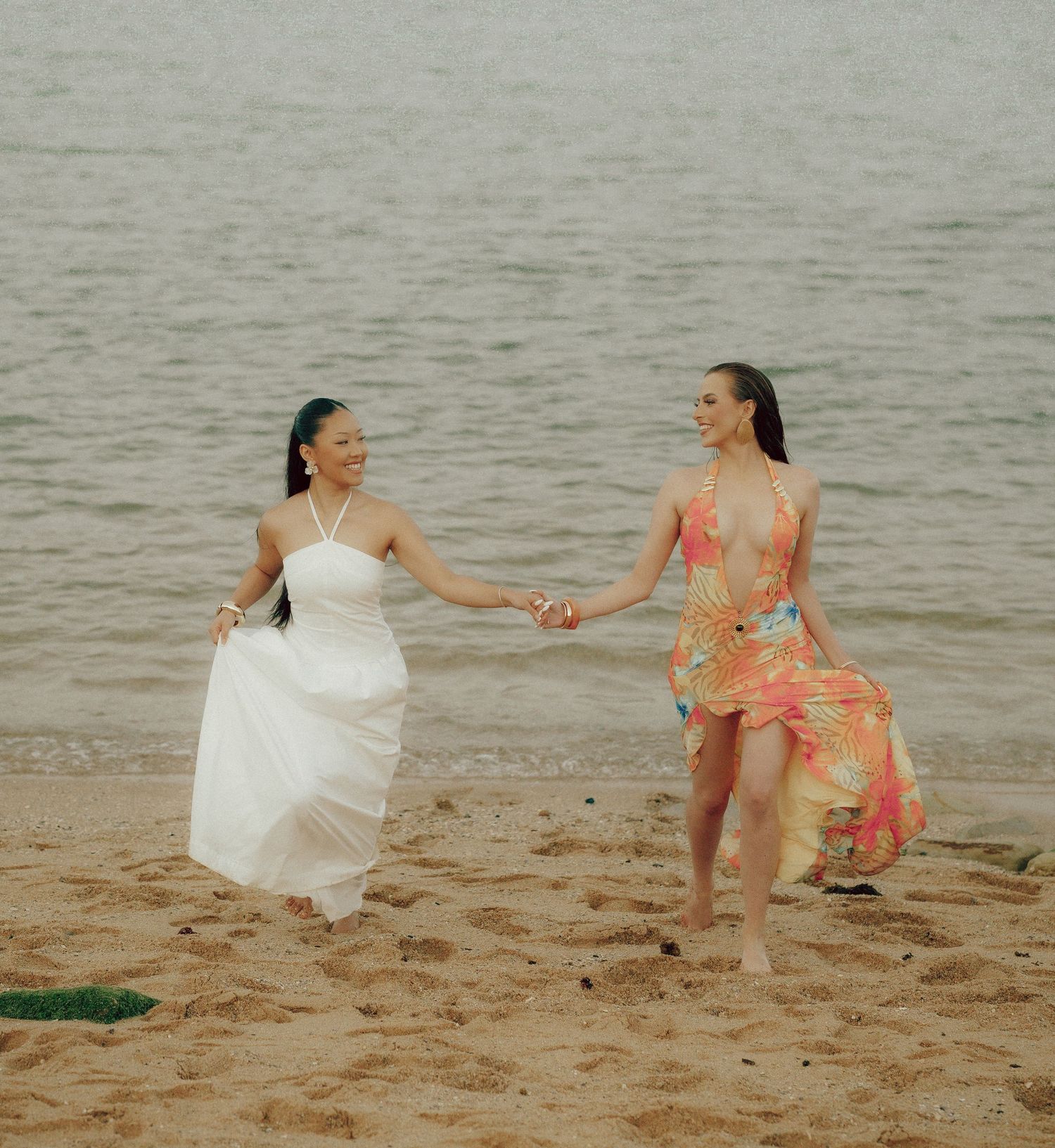 Two smiling women holding hands on a beach shoreline, wearing white and floral summer dresses.