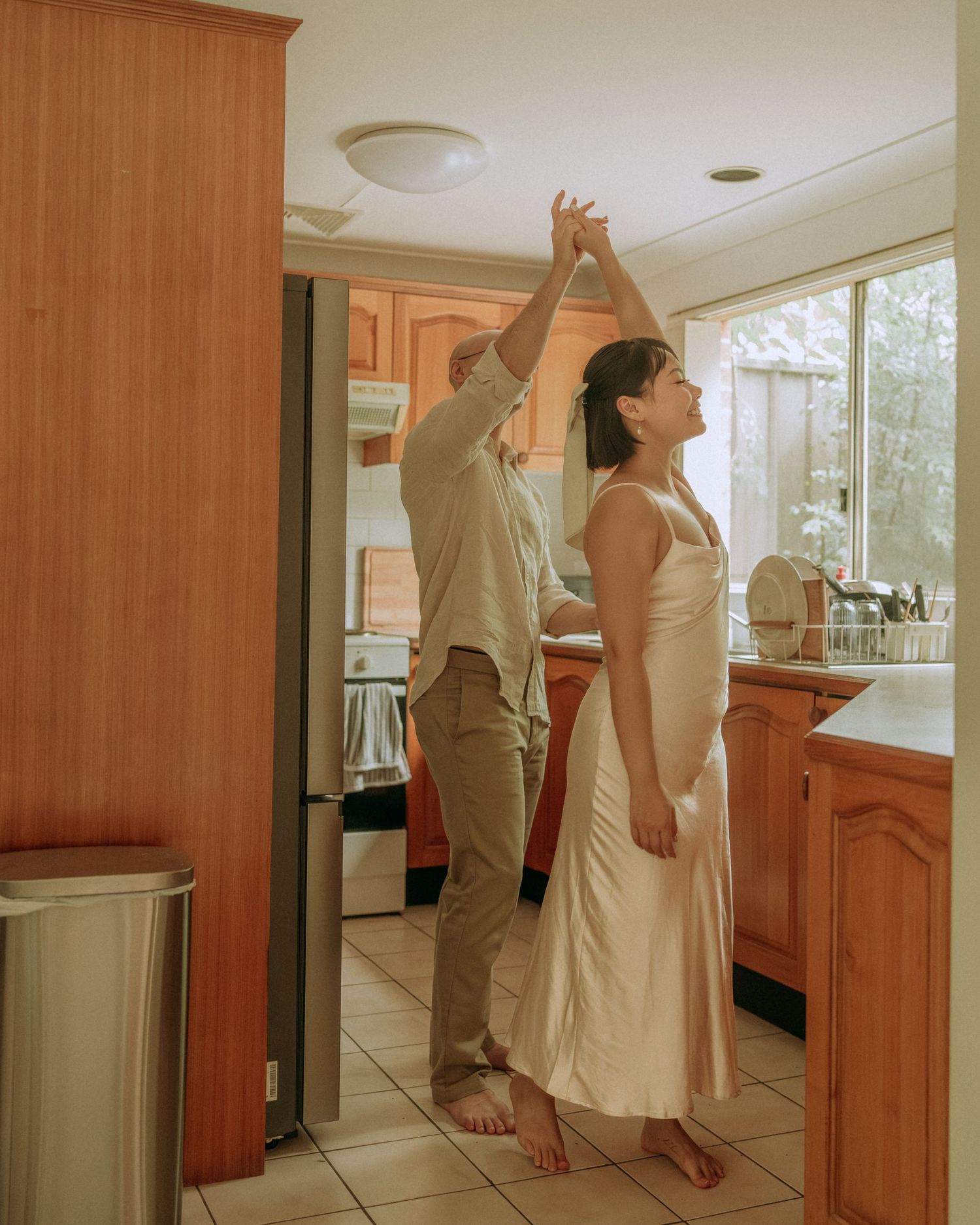 Couple dancing together barefoot in a warm kitchen, man spinning woman in a cream silk slip dress.