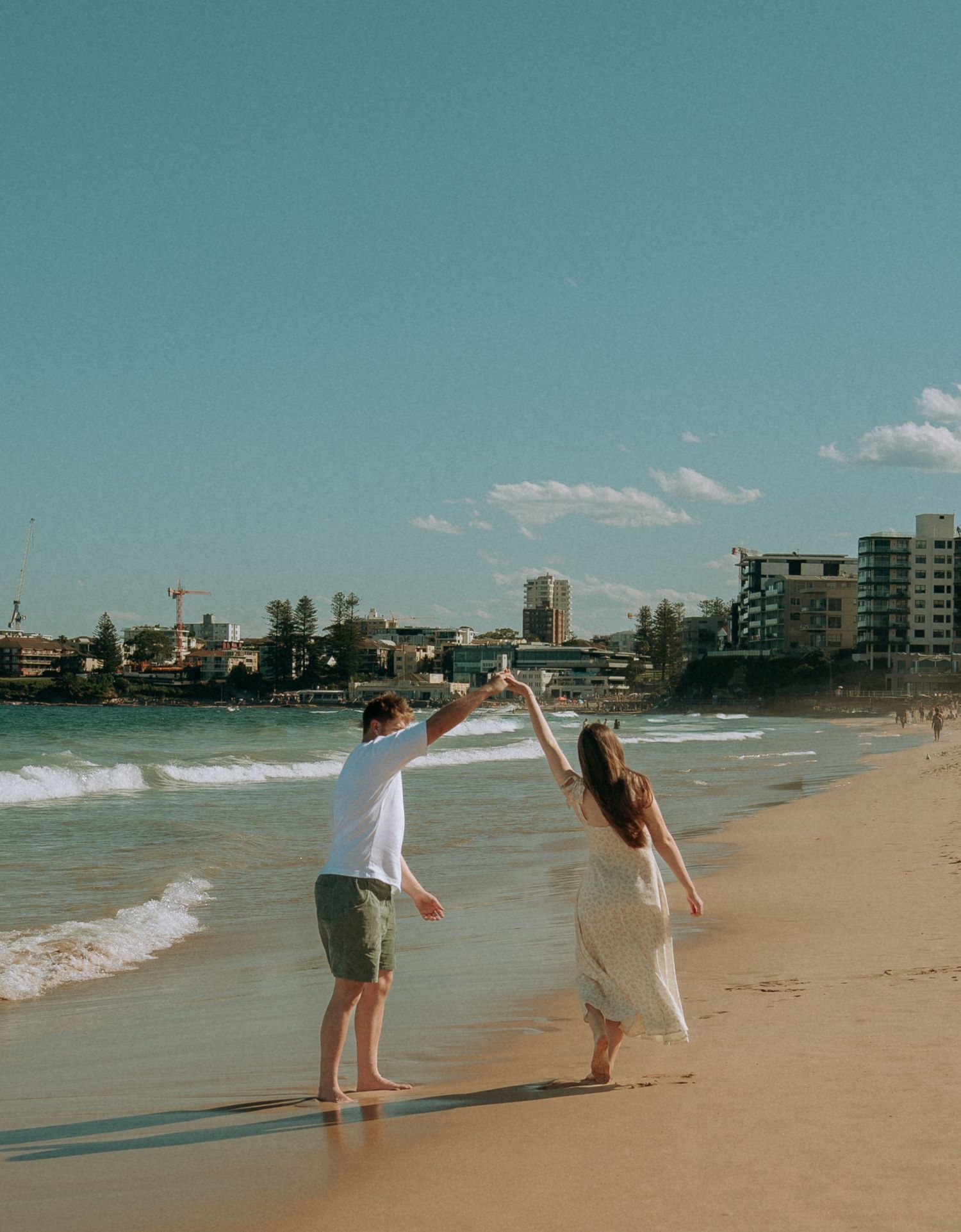 A couple dances on a sandy beach with ocean waves and a coastal cityscape under a clear blue sky.