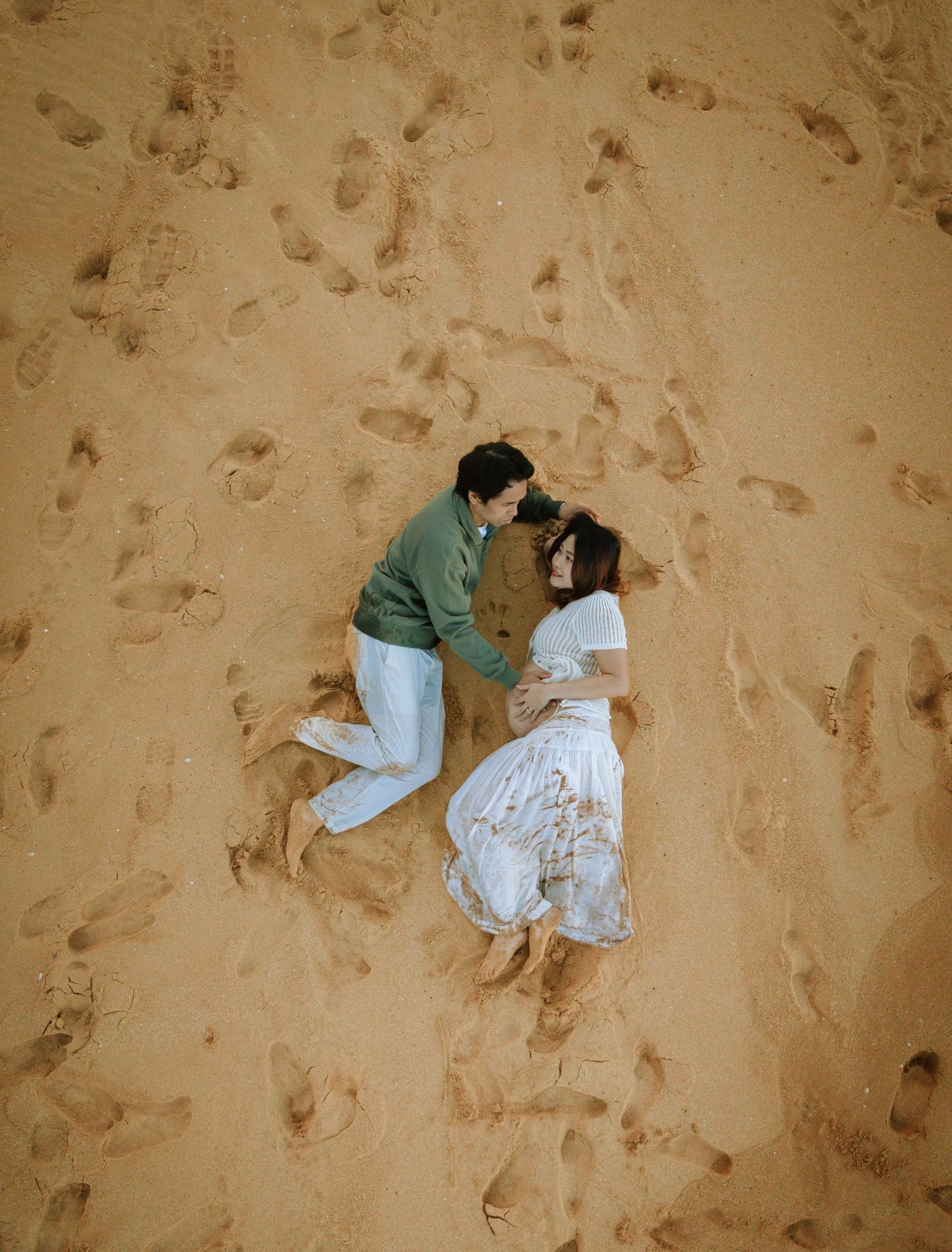 Aerial view of a couple lying on sandy beach surrounded by footprints, woman in white dress, man in green sweater.