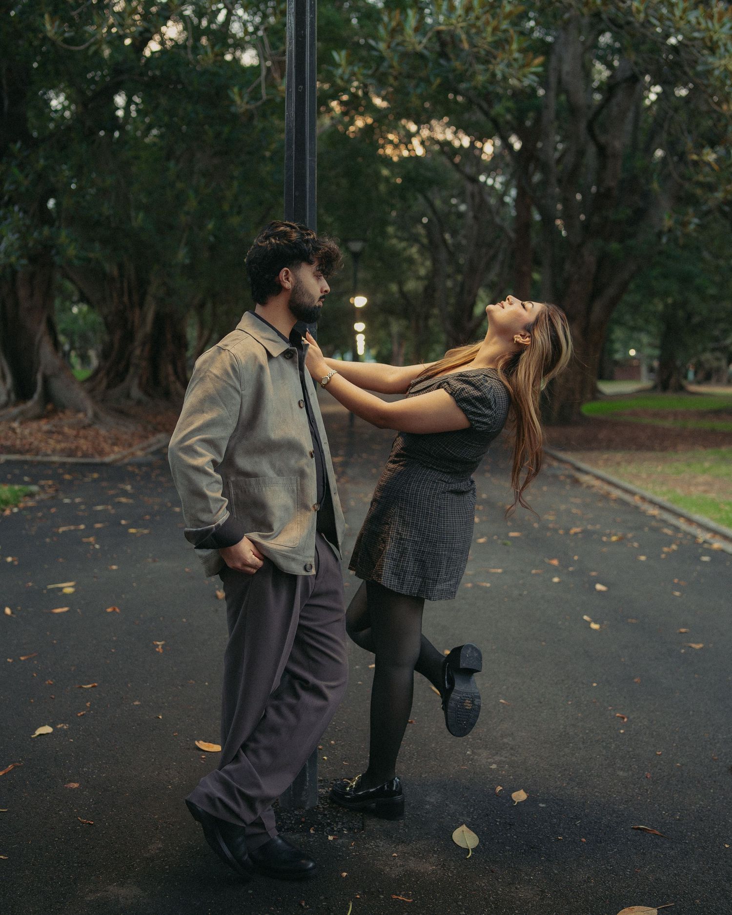 Couple sharing a playful romantic moment on a tree-lined park pathway at dusk.