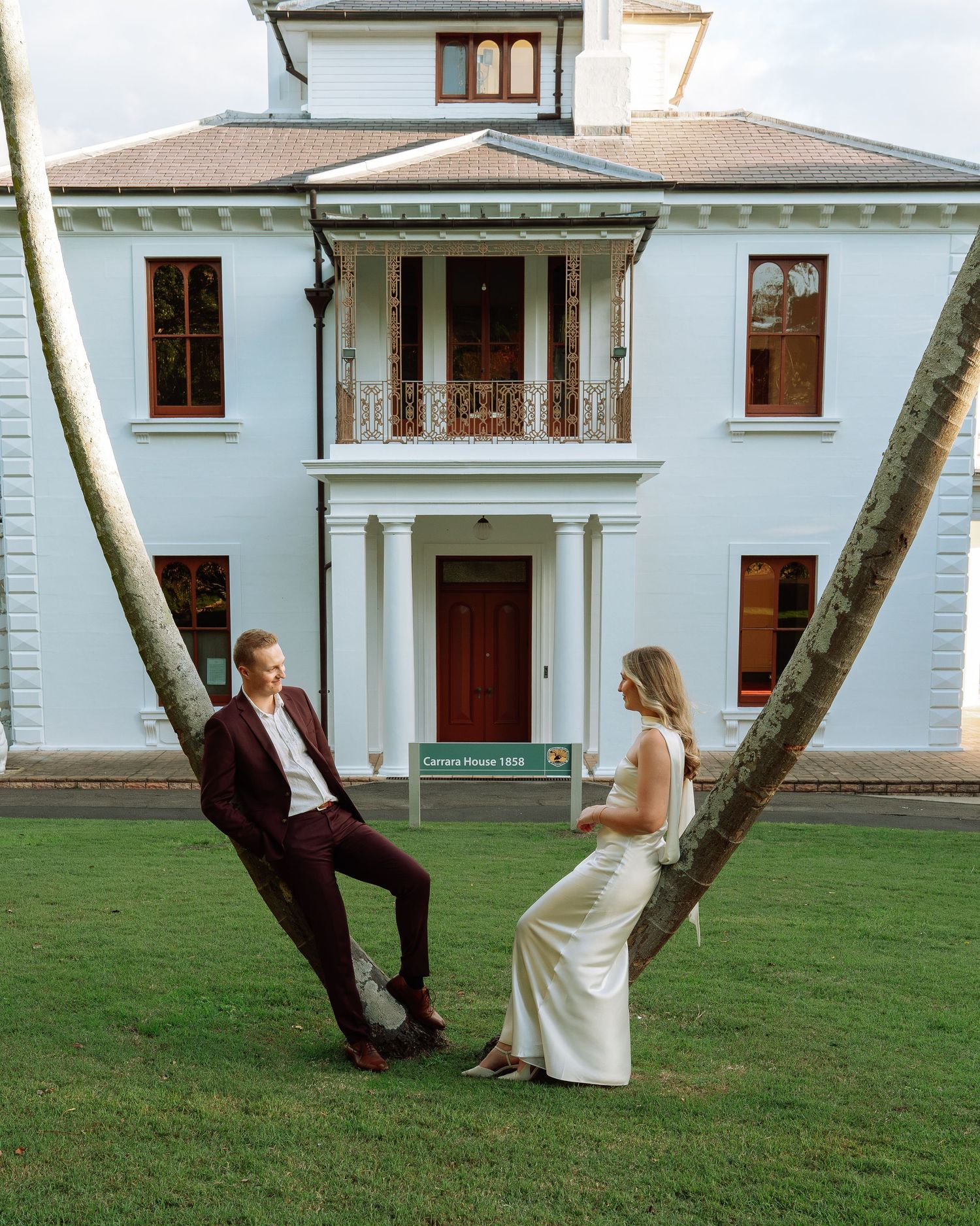 Bride and groom lean against crossed palm trees in front of historic white colonial building.