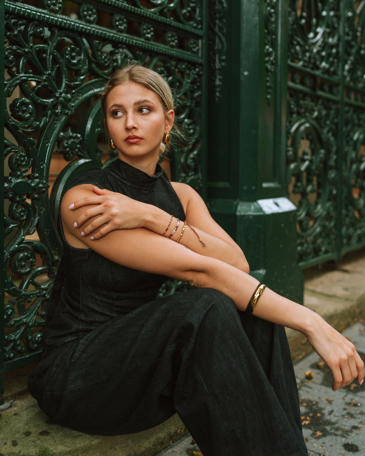 Woman in black sleeveless dress sitting against ornate green iron gate, wearing gold jewelry.