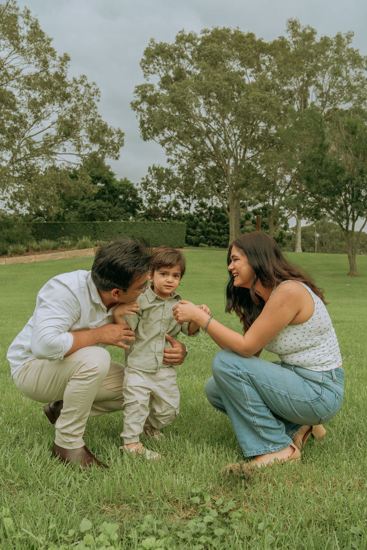 Parents crouching down to interact with their smiling toddler on green grass.