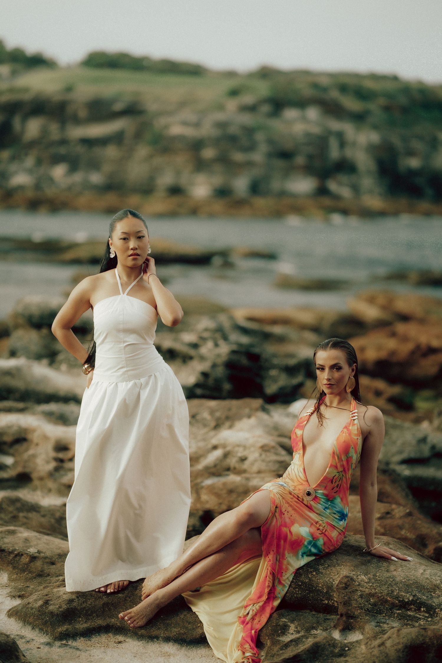 Two women in white and floral dresses posing on rocky seaside terrain at dusk.