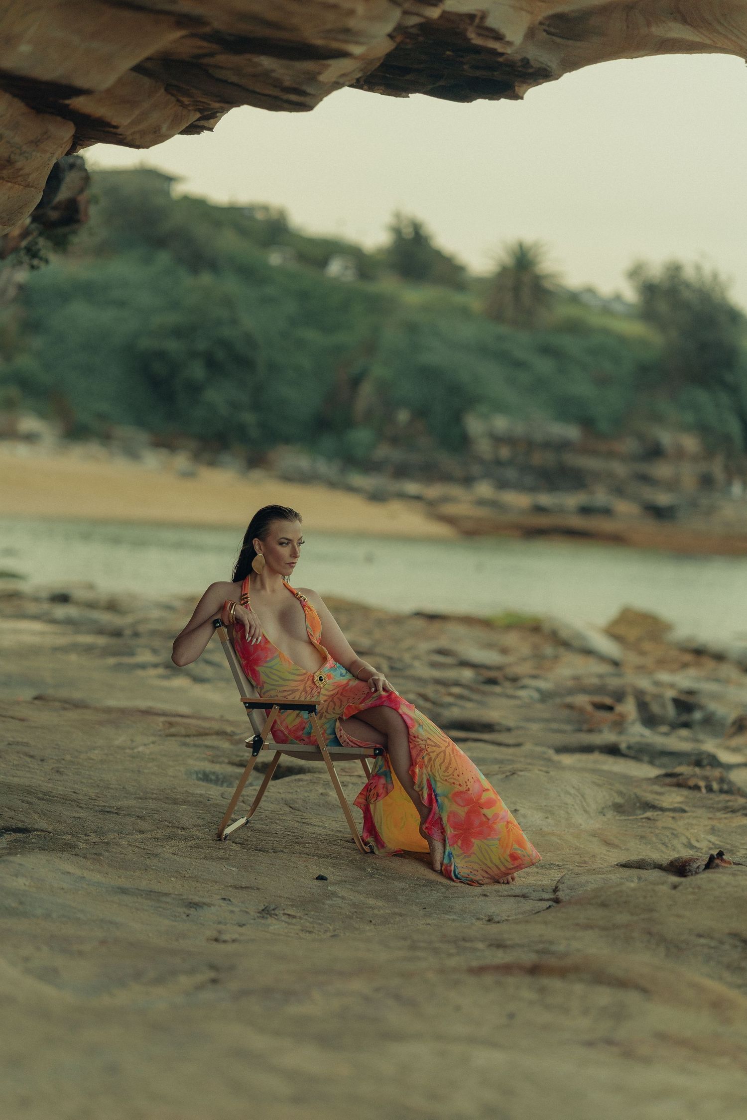 Woman in colorful floral maxi dress sitting on chair among coastal rocks with beach and greenery backdrop.