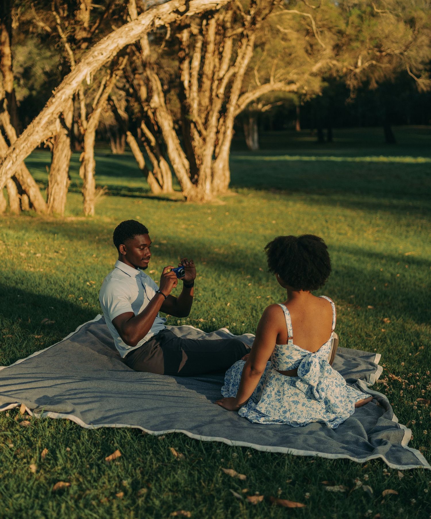 A couple relaxes on a picnic blanket in a tree-lined park, sharing a peaceful golden hour moment.