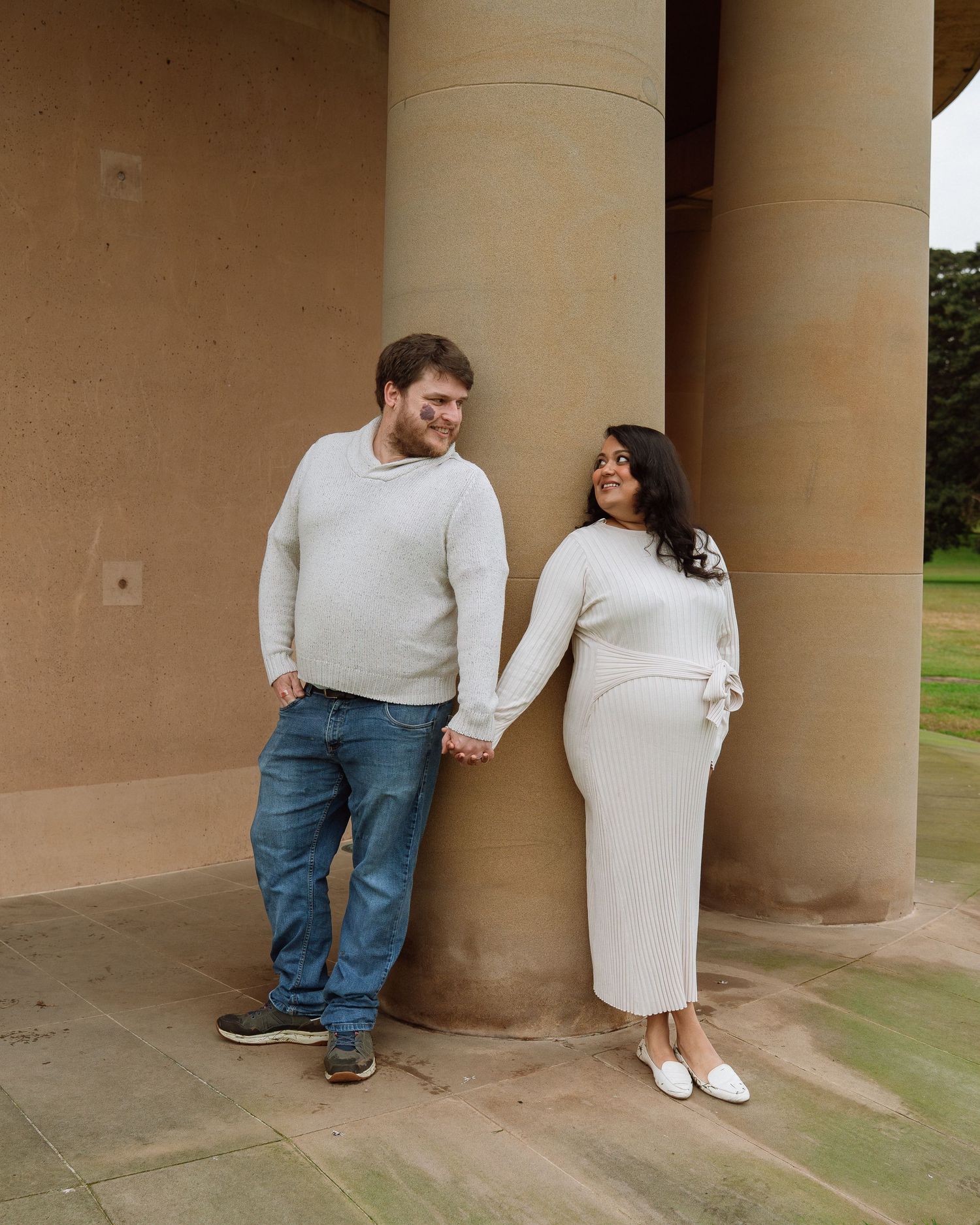 Expecting couple holding hands and smiling beside large columns during an outdoor maternity shoot.