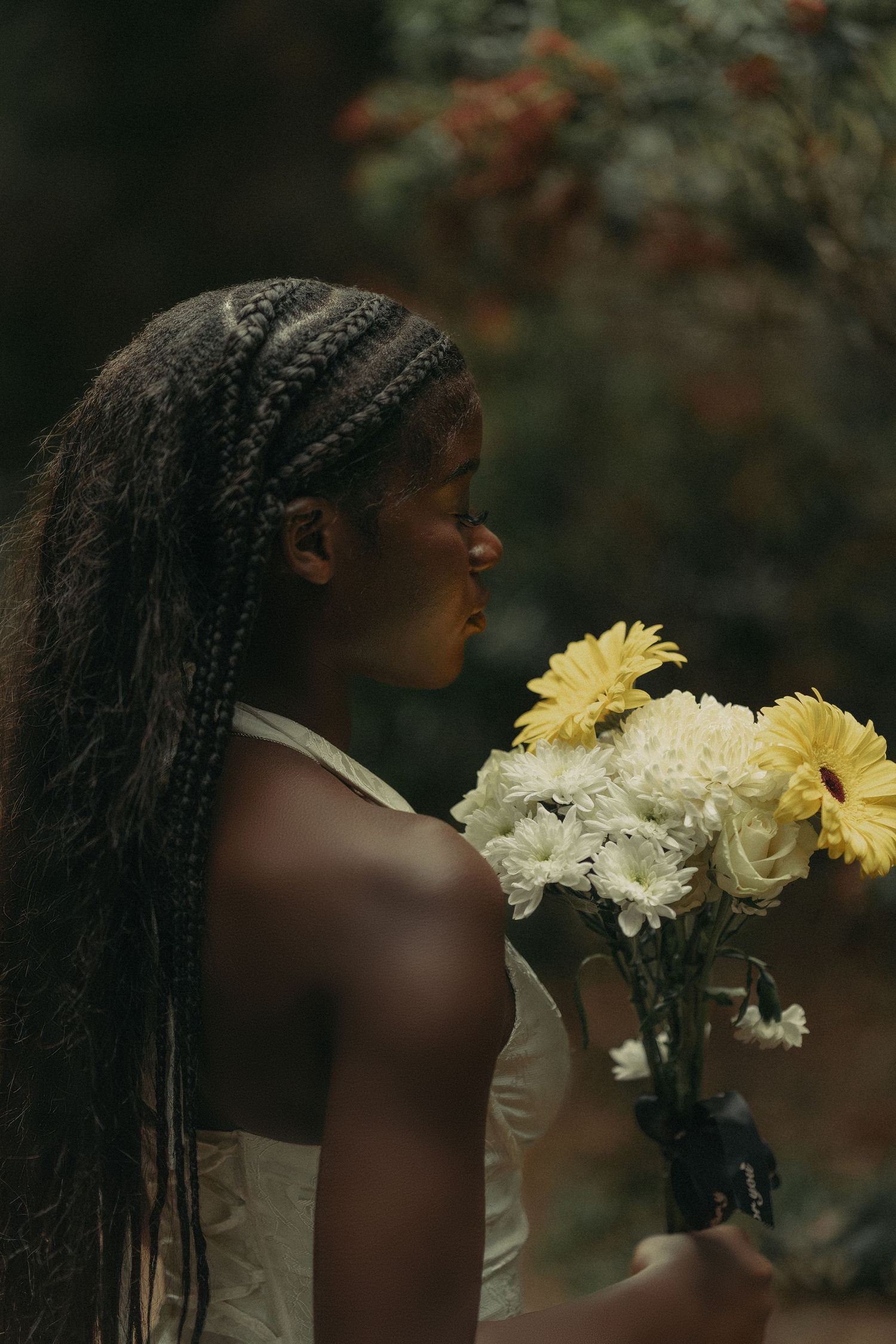 Young Black woman with braids holding yellow and white flowers in dark garden setting.