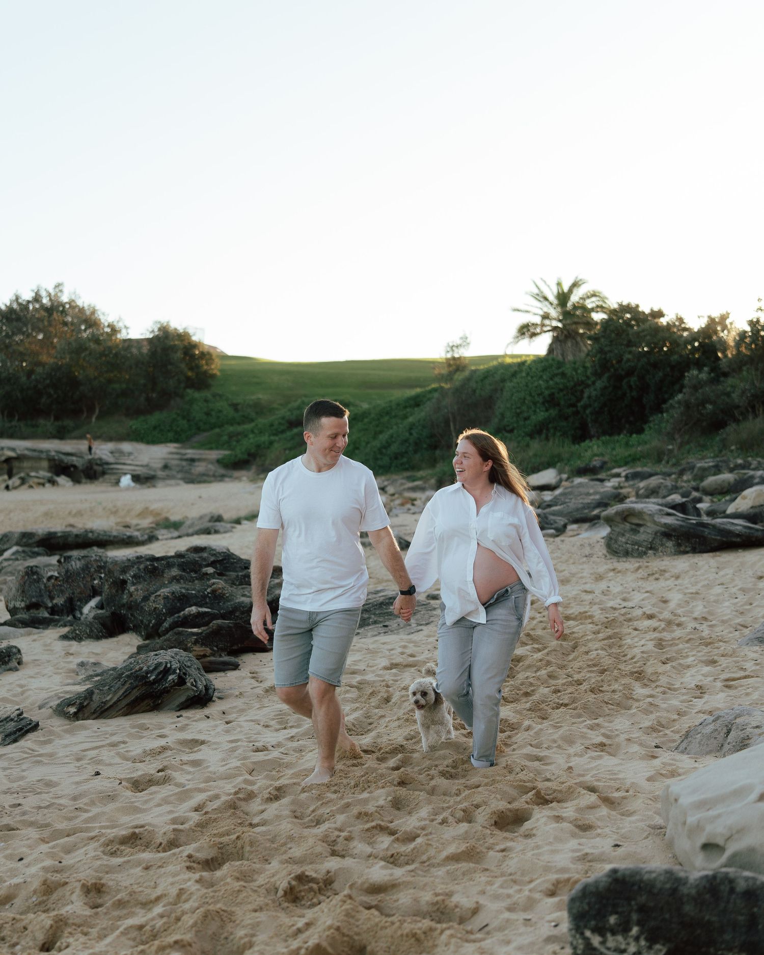 Pregnant couple walking hand in hand on sandy beach with small dog, lush greenery in background.