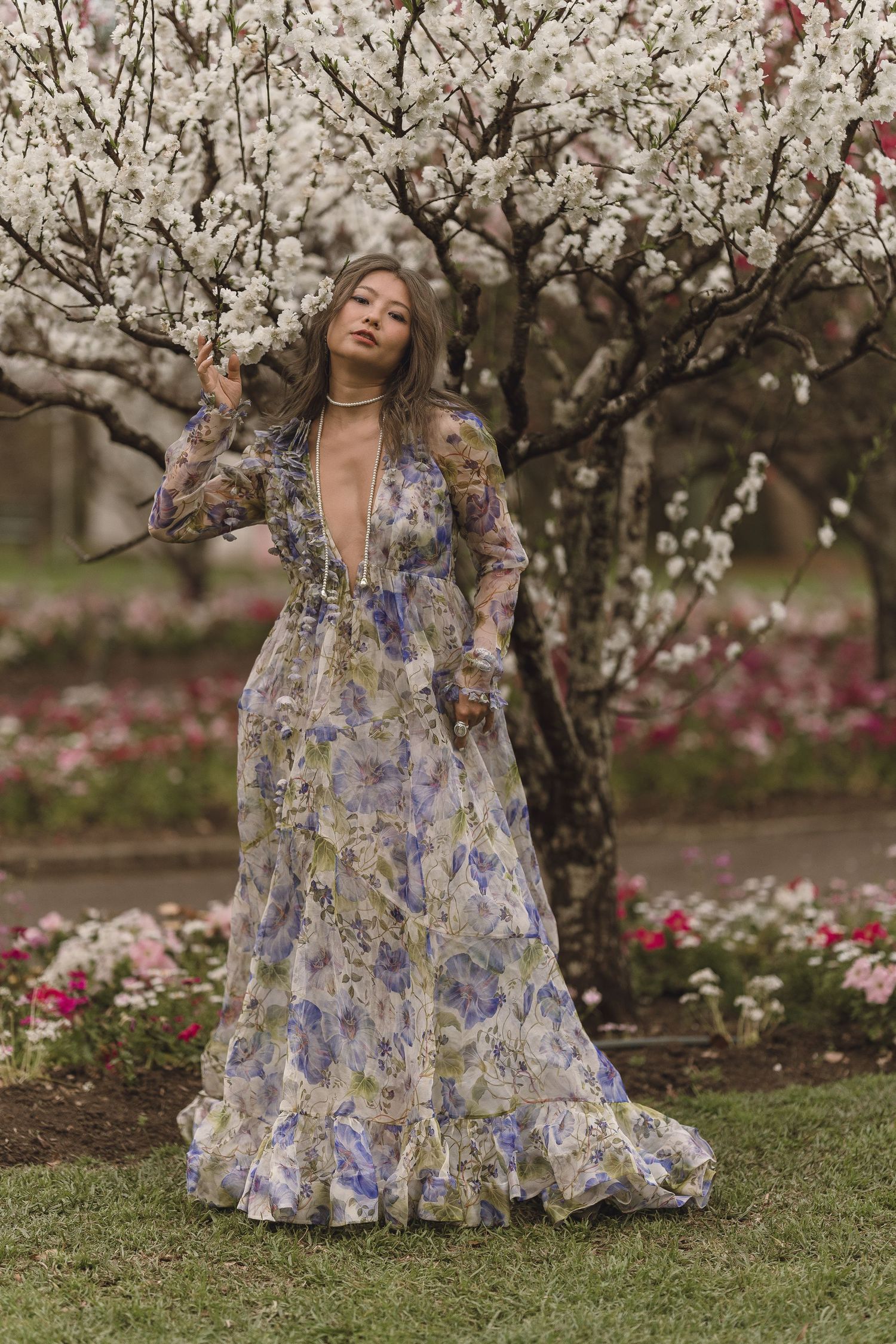 Asian woman in deep-V floral maxi dress posing beside white cherry blossom tree in colorful garden.