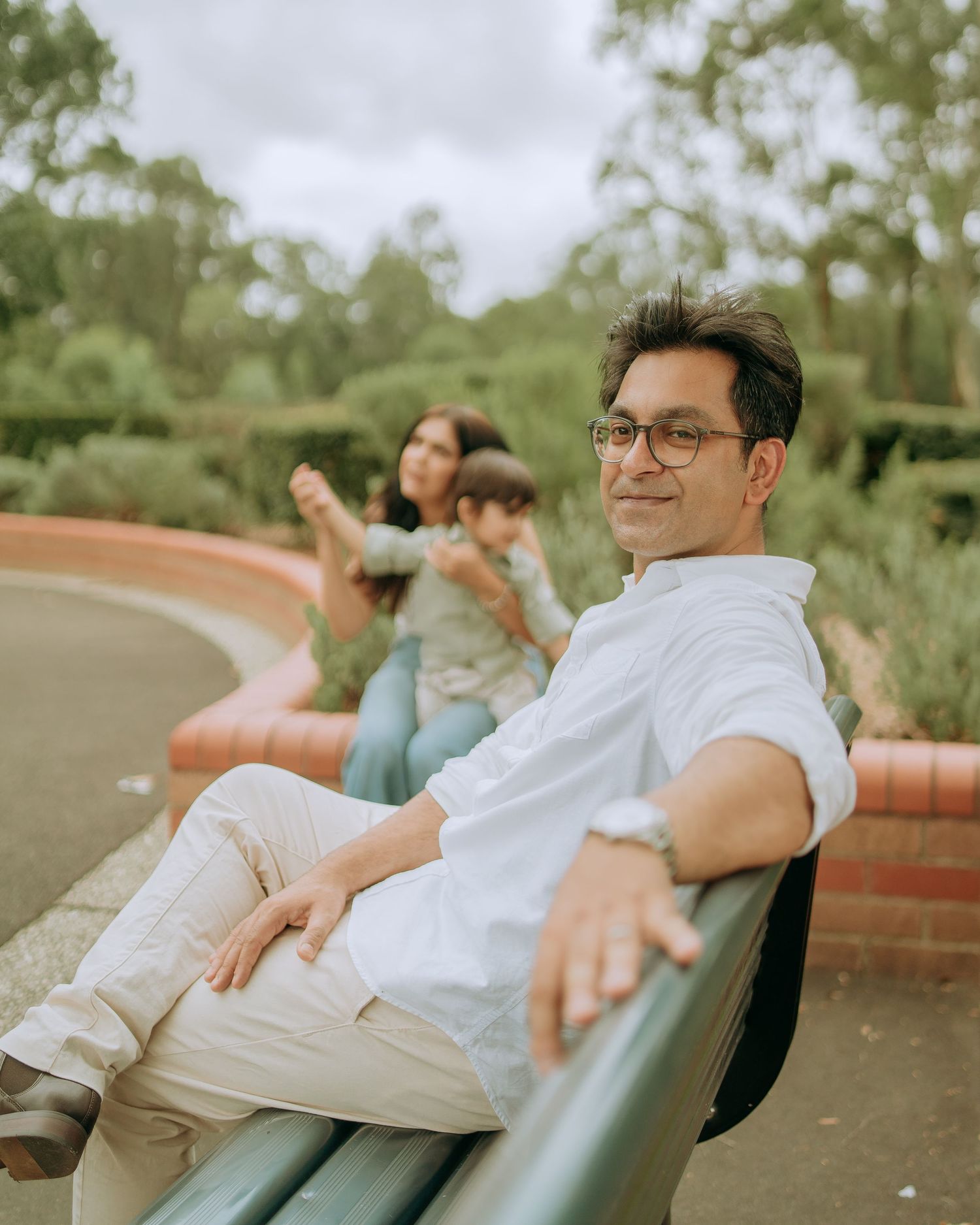 Man in white shirt relaxing on park bench smiling at camera while family plays in background.