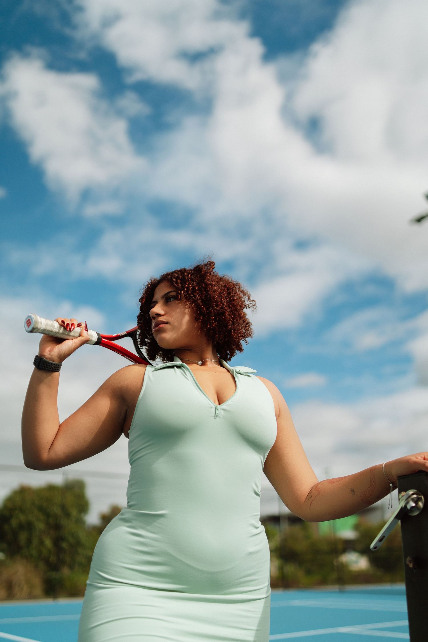 A curly-haired woman in a mint green tennis dress holds a red racket on her shoulder at an outdoor blue court.