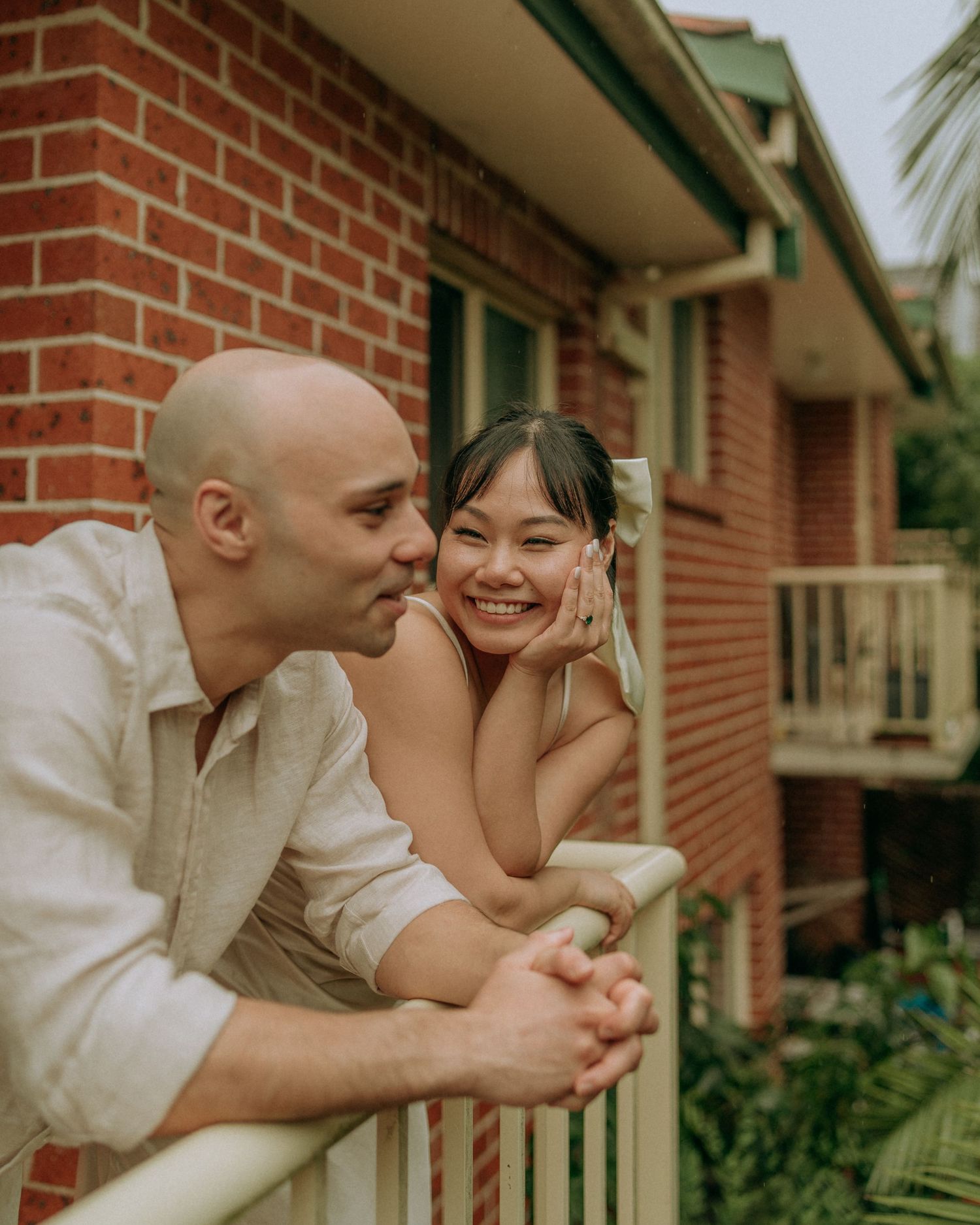 Smiling couple leaning on white balcony railing of brick apartment building outdoors.