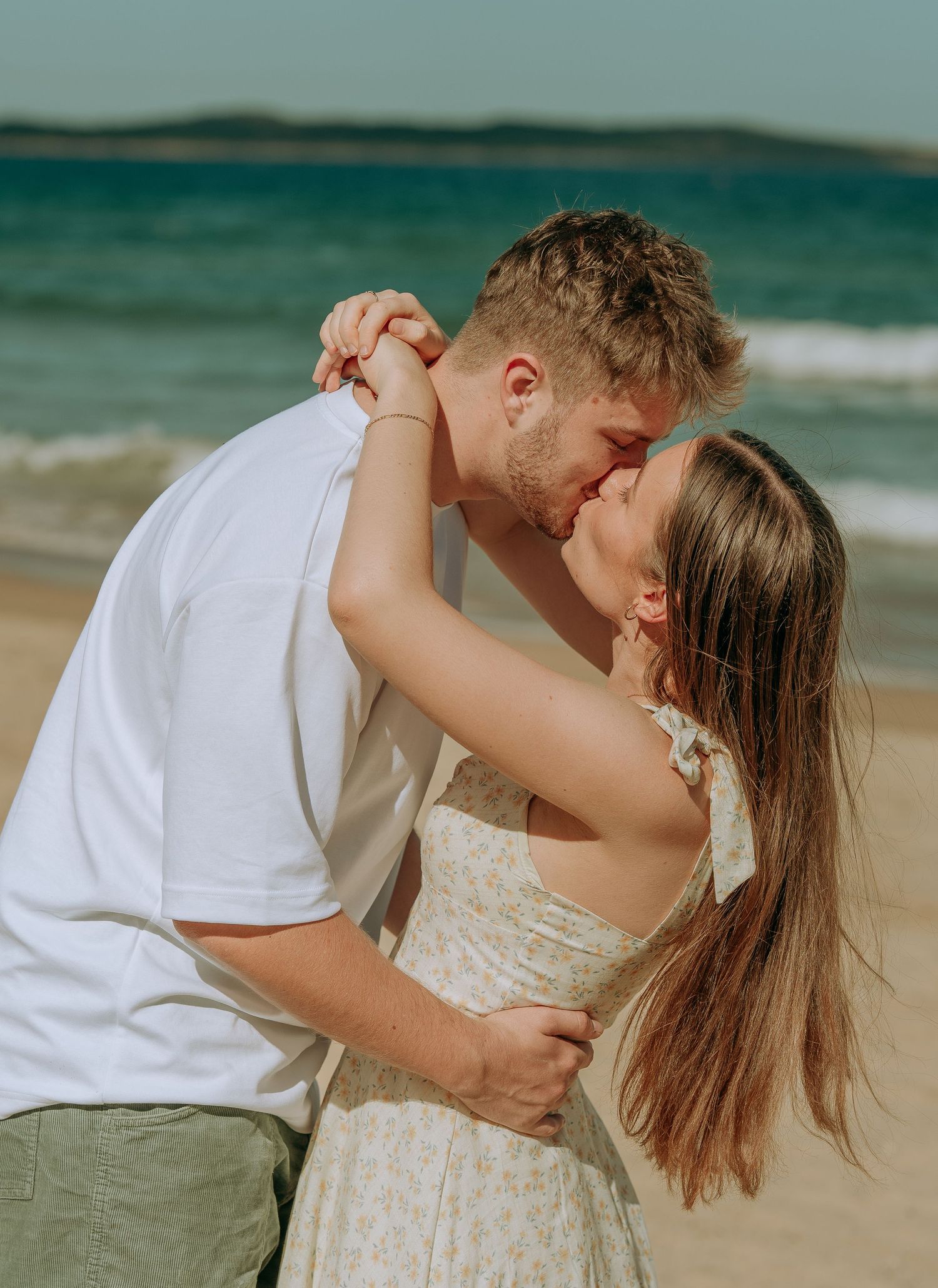 Couple sharing romantic kiss on sandy beach, woman in floral dress, man in white tee and green shorts.