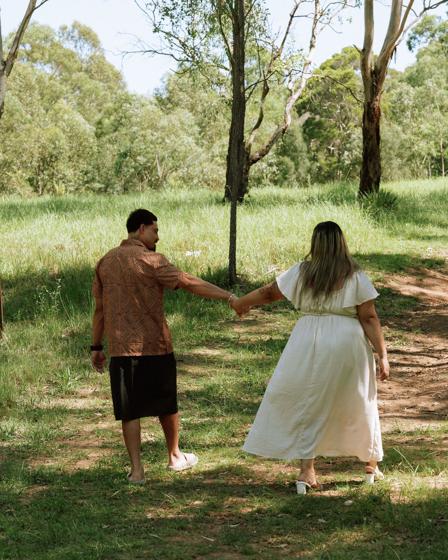 Couple walking hand in hand along a dirt path through a vibrant green Australian bushland landscape.