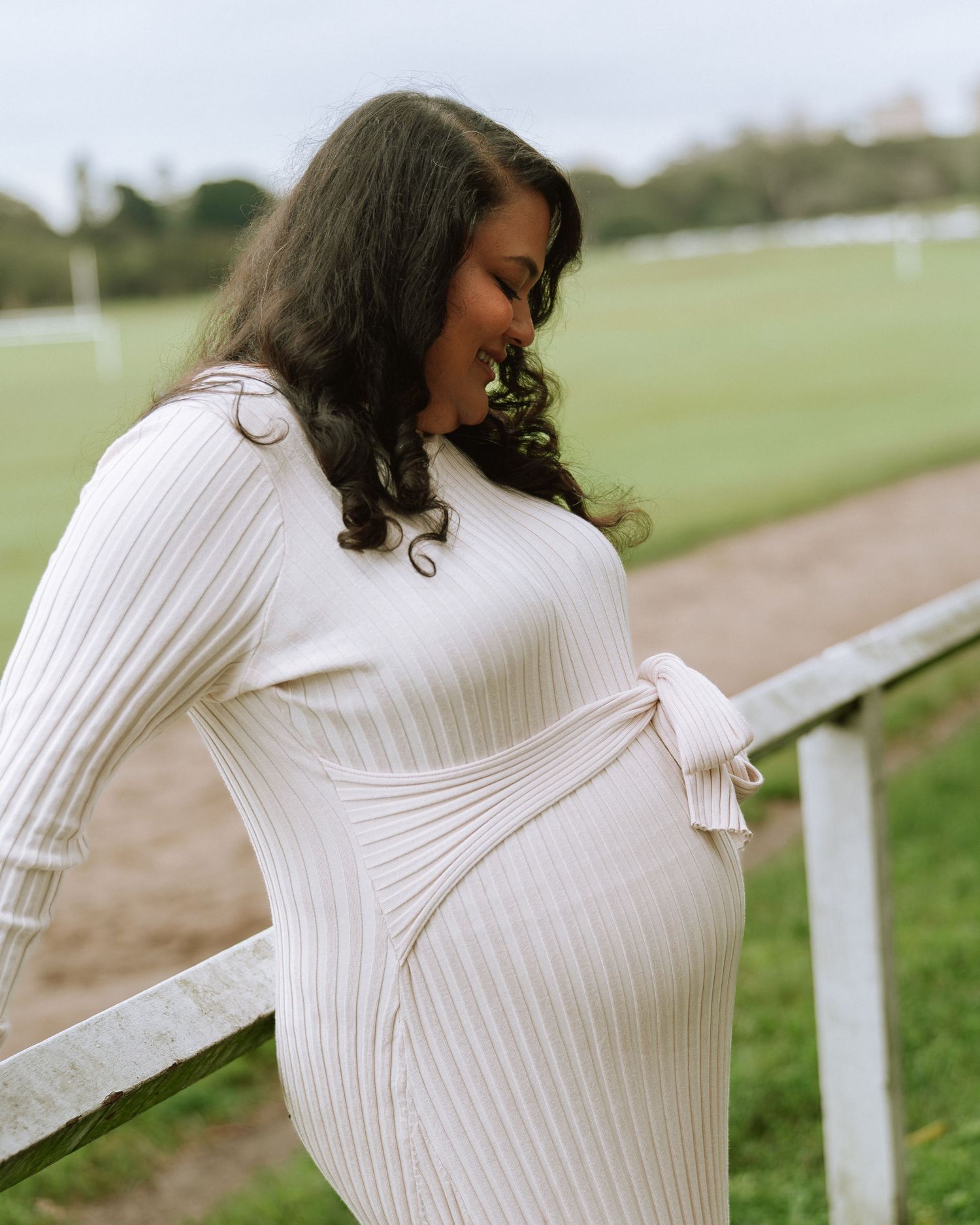 Pregnant woman in white ribbed maternity dress leaning on a fence, smiling at her baby bump.