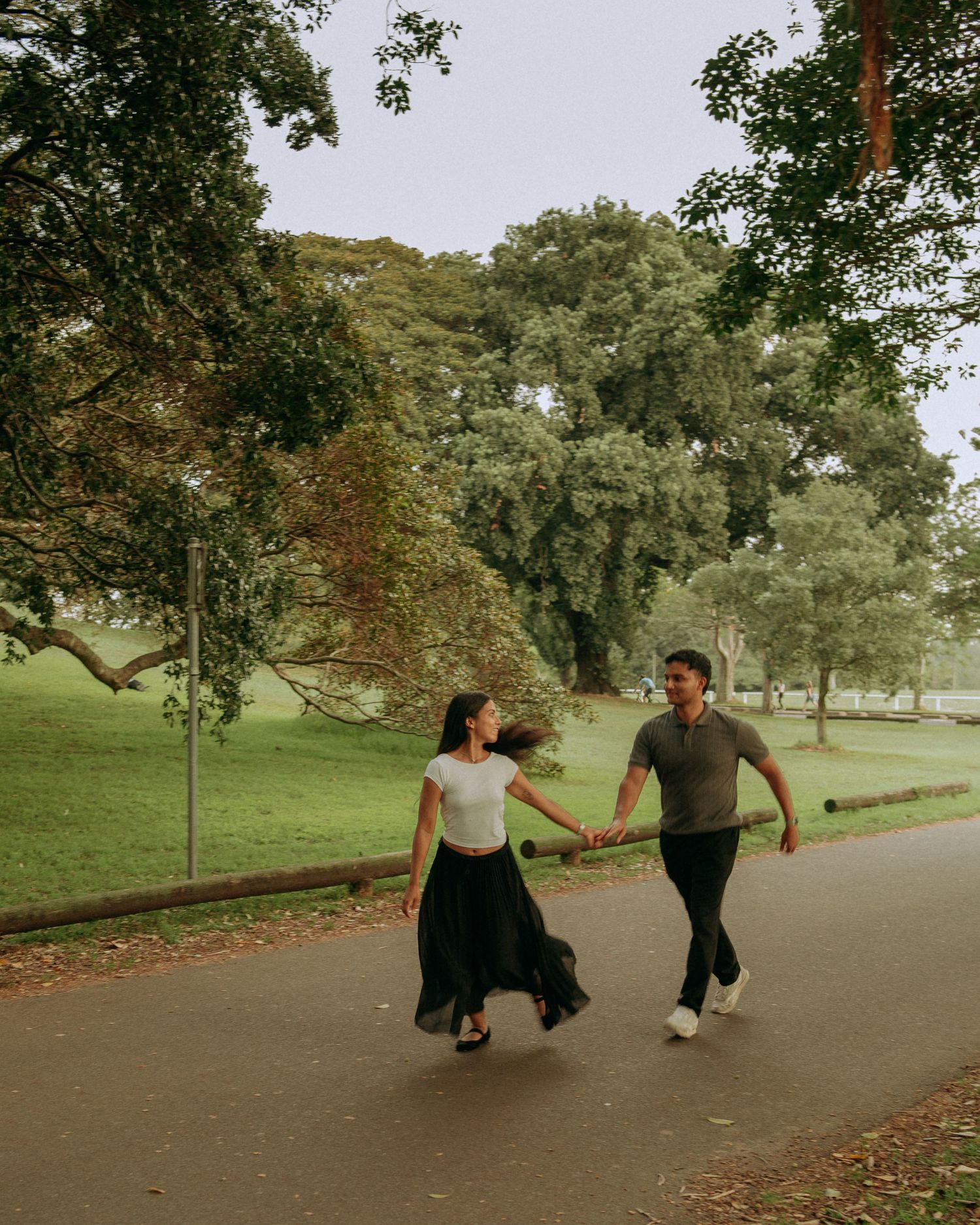 Couple holding hands and walking joyfully along a tree-lined park path.
