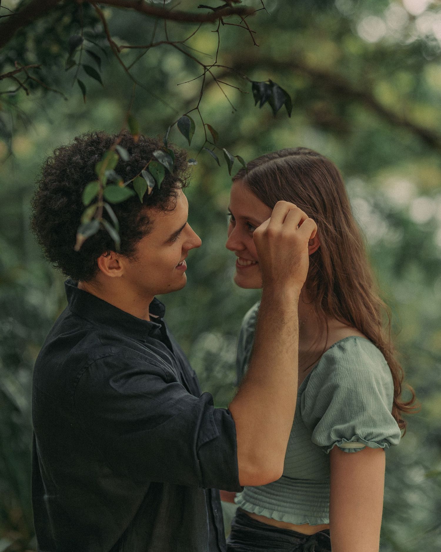 A couple shares a tender moment outdoors, the man gently touching the womans face amid lush green trees.