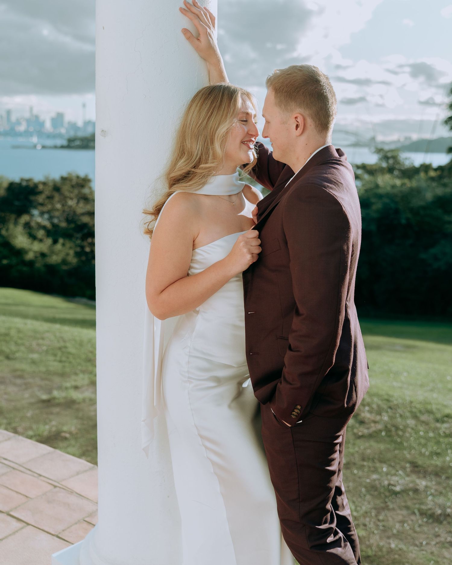 Bride in white jumpsuit and groom in burgundy suit share romantic moment leaning against white column outdoors.