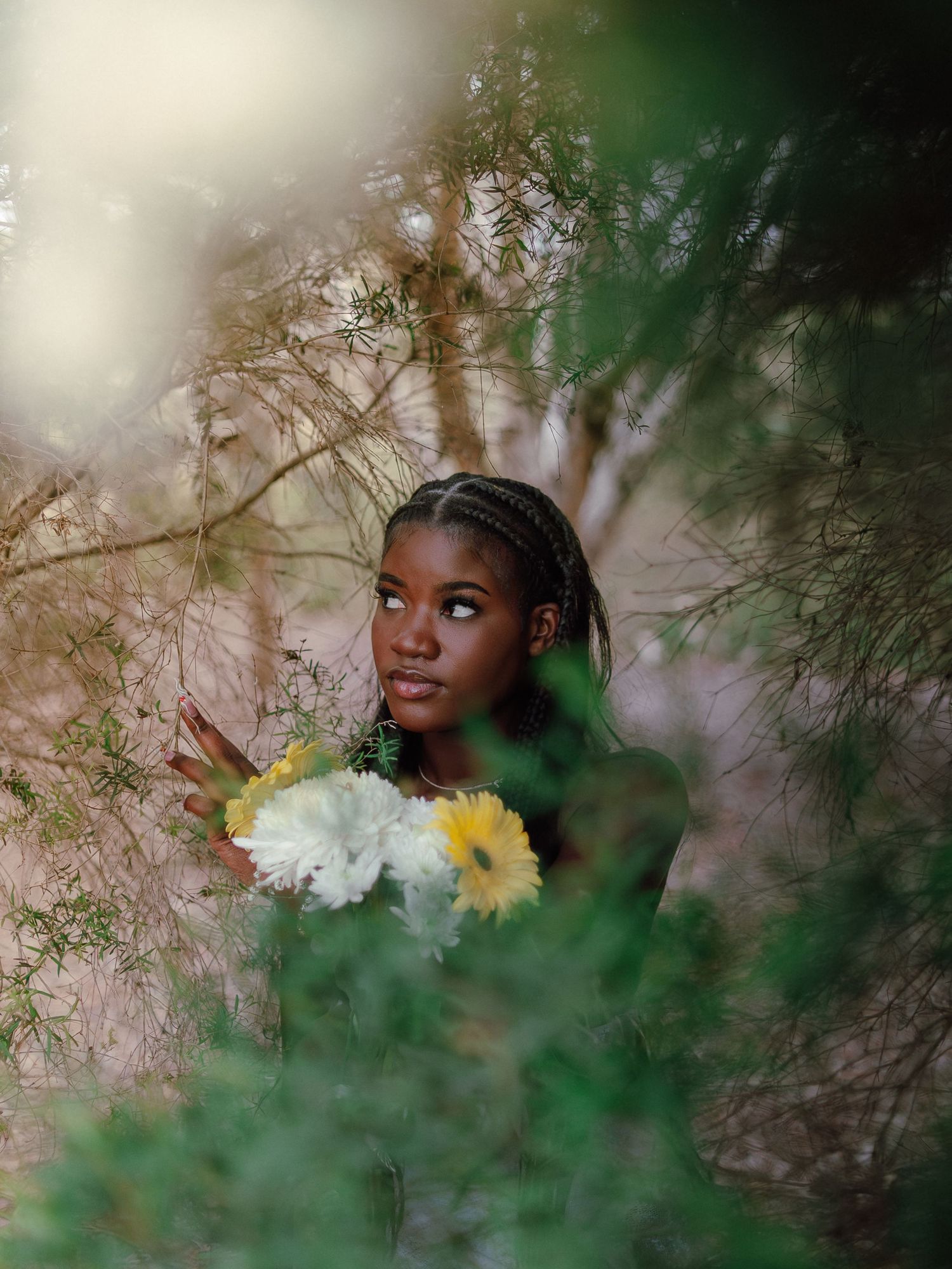 Young Black woman with braids holds colorful flowers while peeking through lush green foliage.