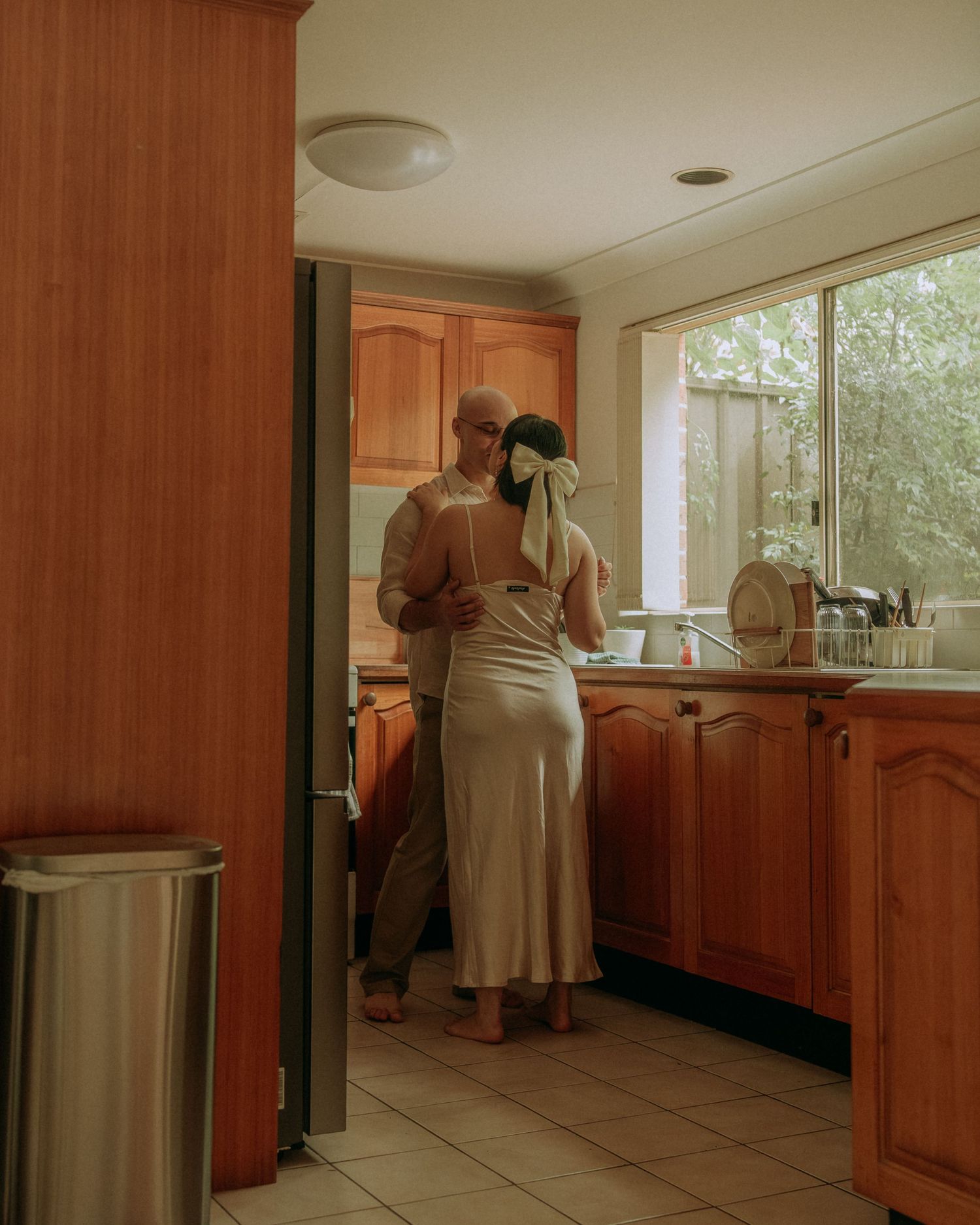 Couple embracing and kissing in a warm wooden kitchen viewed from doorway in soft natural light.