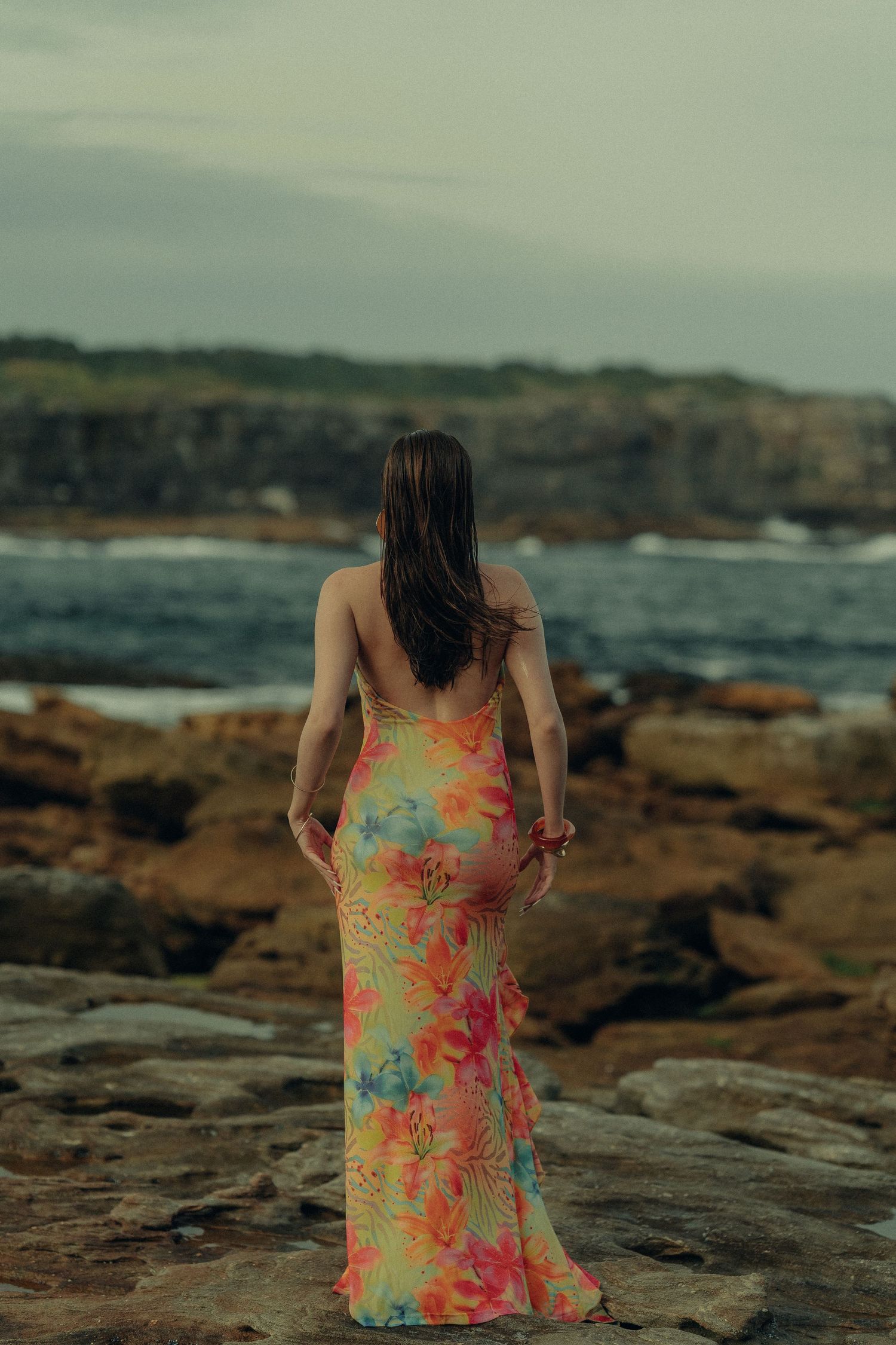 Woman in bright floral maxi dress standing on rocky coastal cliffs overlooking crashing ocean waves.