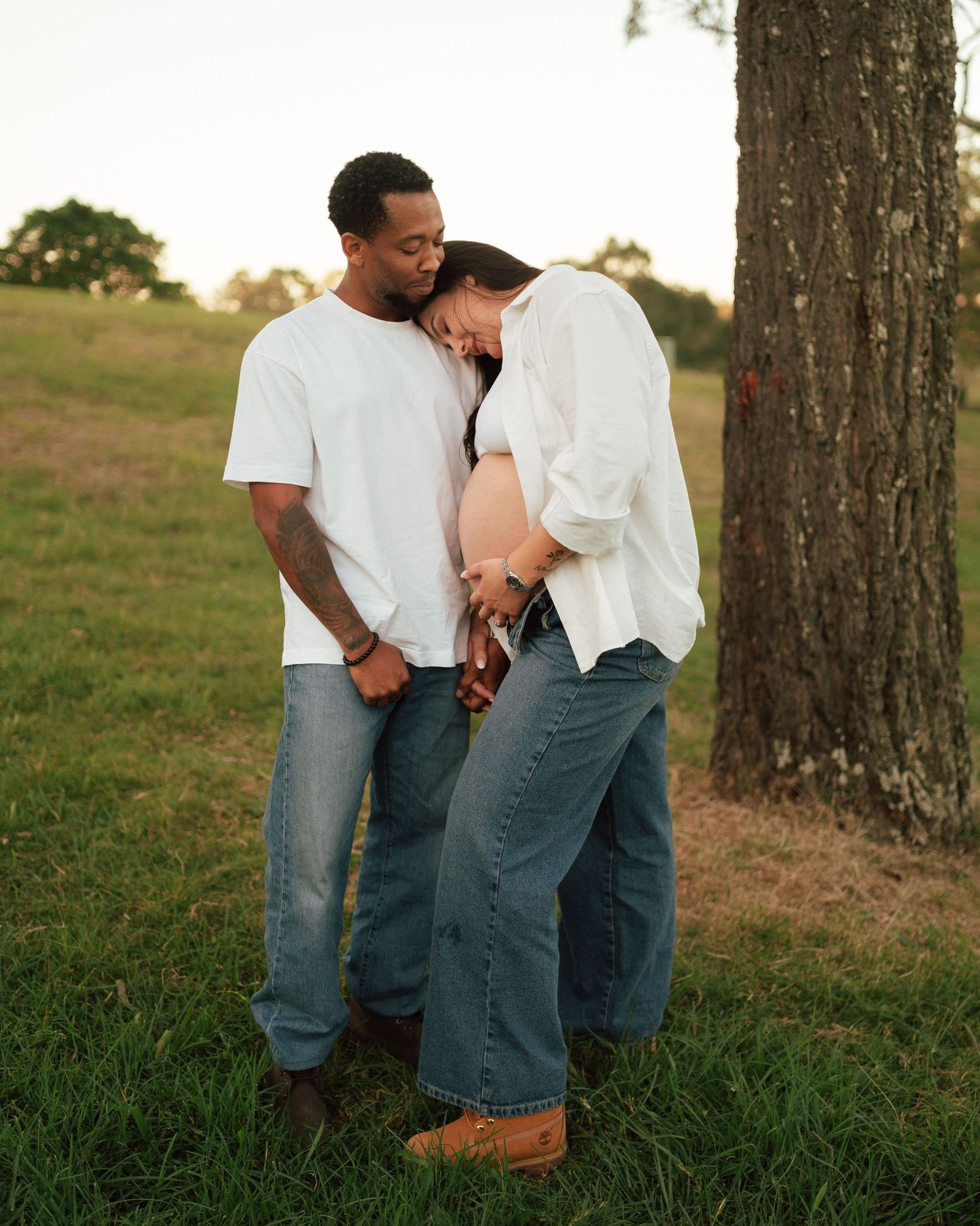 Couple standing by a tree at sunset, pregnant woman leaning on partners shoulder holding hands tenderly.