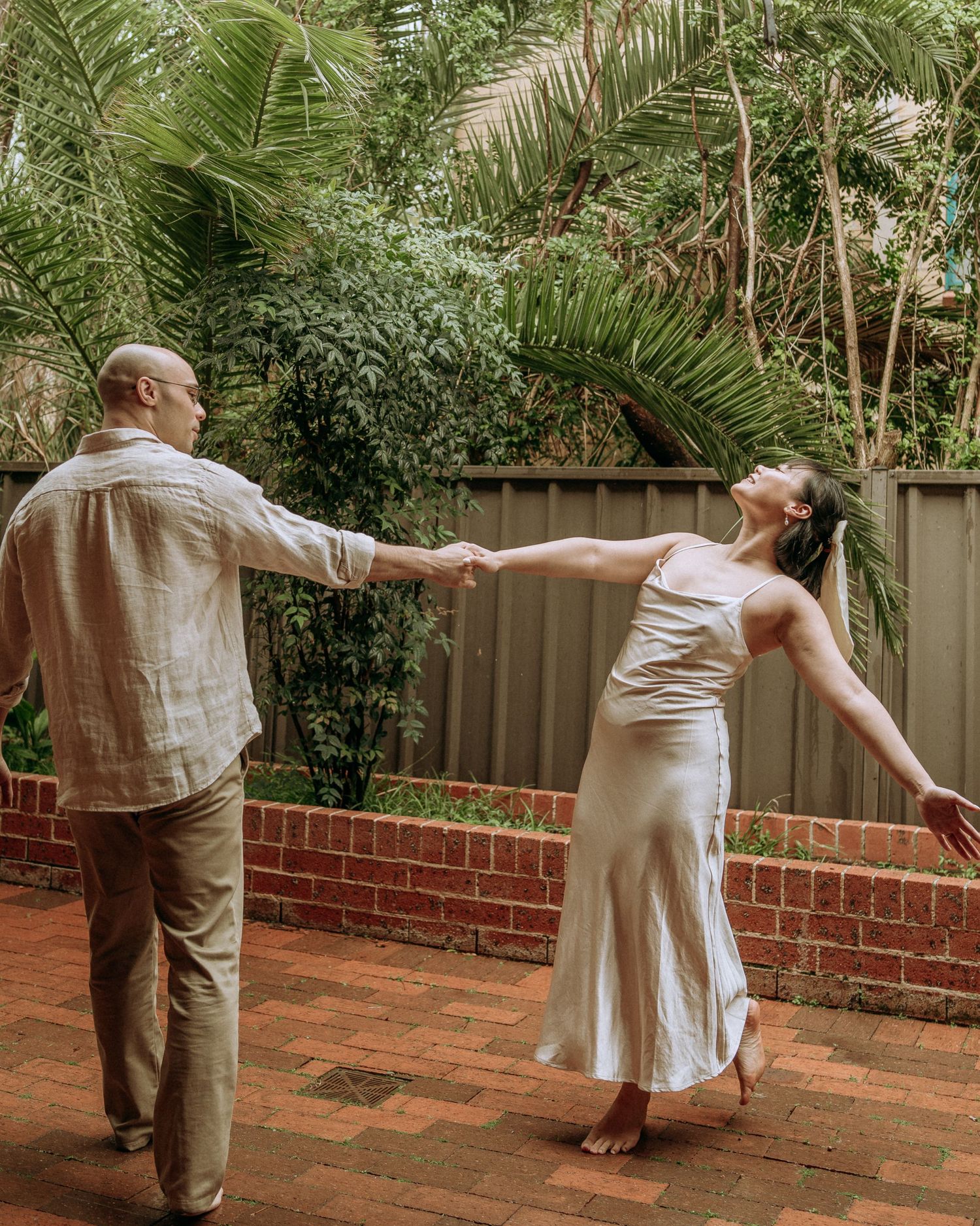 Couple dancing barefoot on brick patio surrounded by lush tropical garden greenery.