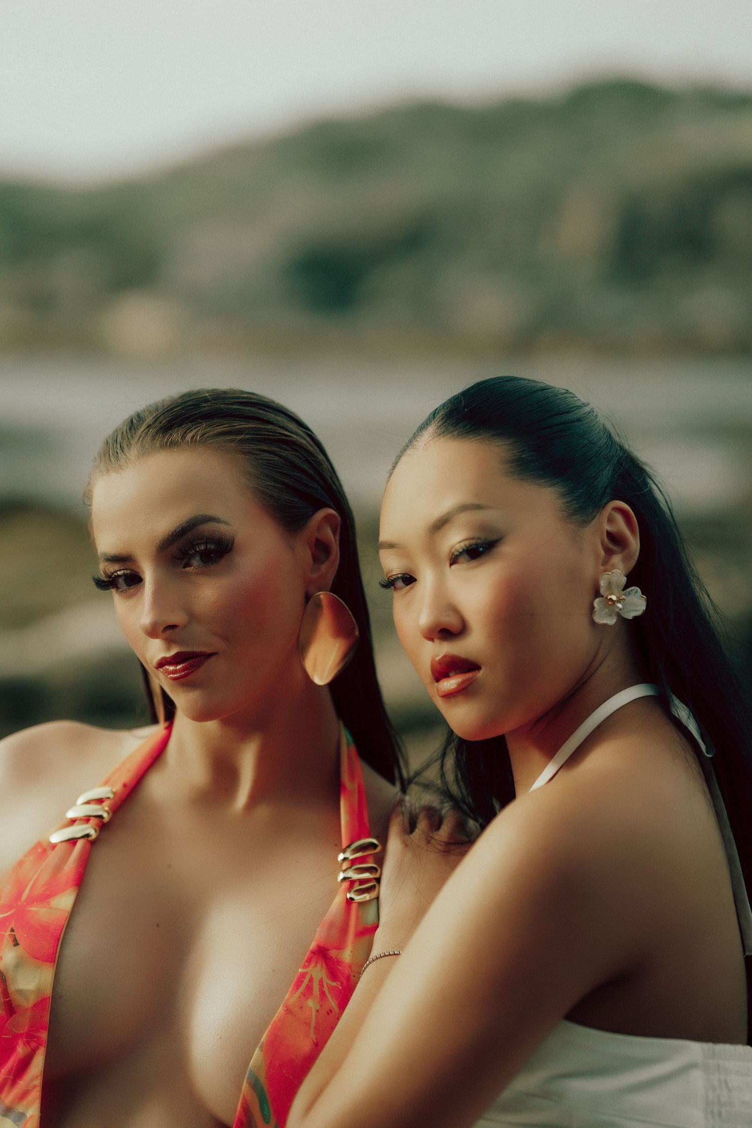 Two glamorous women in colorful swimwear posing closely together at a rocky beach.