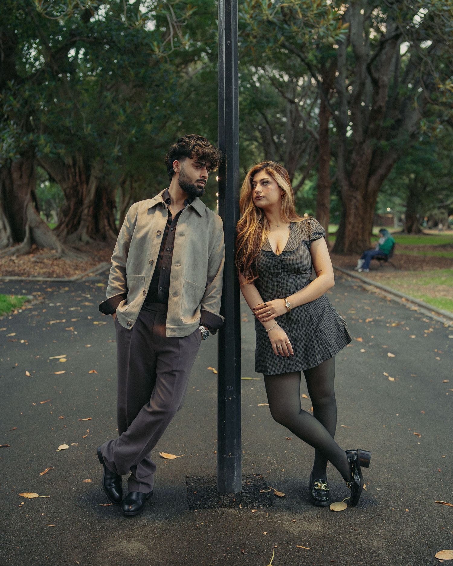 Stylish couple leaning on opposite sides of a lamp post in a tree-lined park pathway.