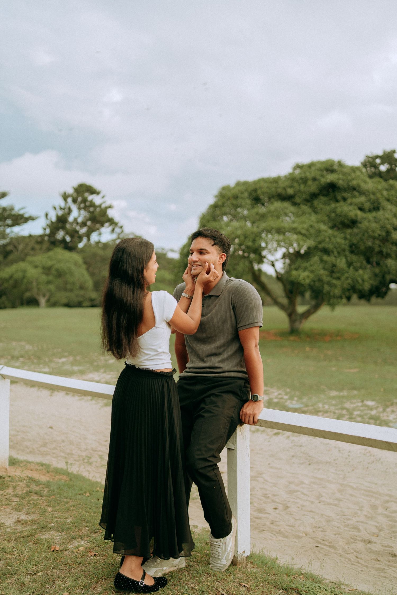 A woman gently cups her smiling partner's face near a white fence in a scenic green park landscape.