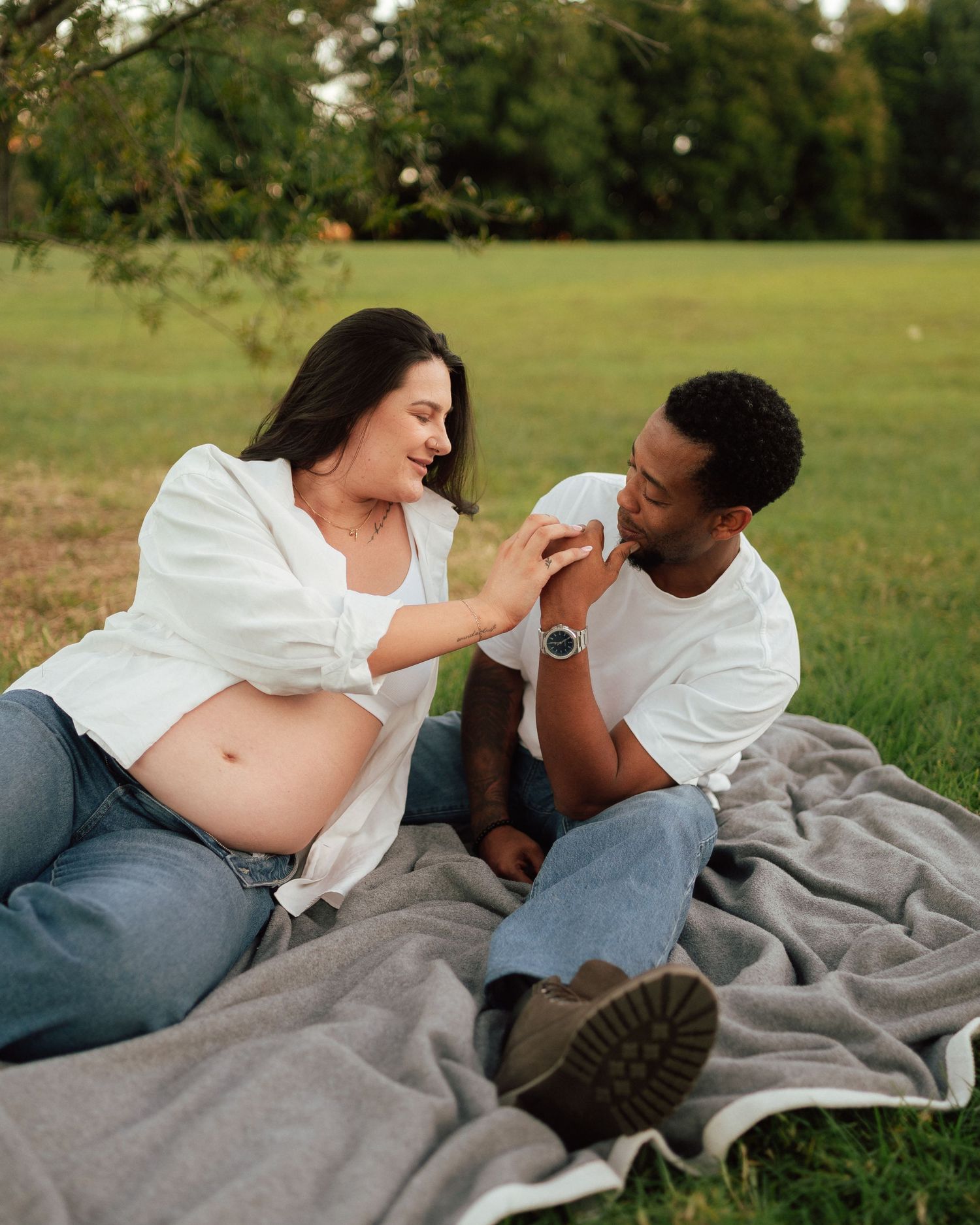 Pregnant couple sitting on a blanket in a park at golden hour, holding hands and smiling at each other.