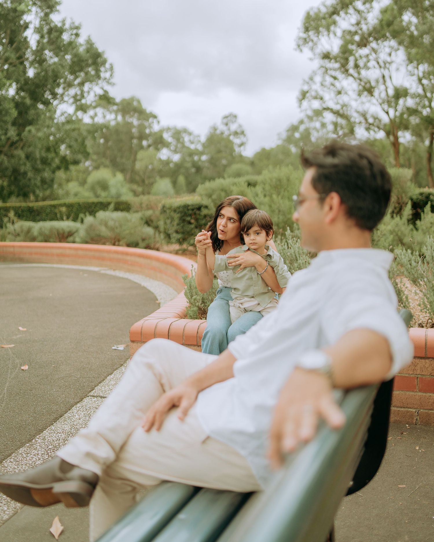 A mother blows bubbles while holding her toddler on a bench as the father sits relaxed in the foreground at a park.
