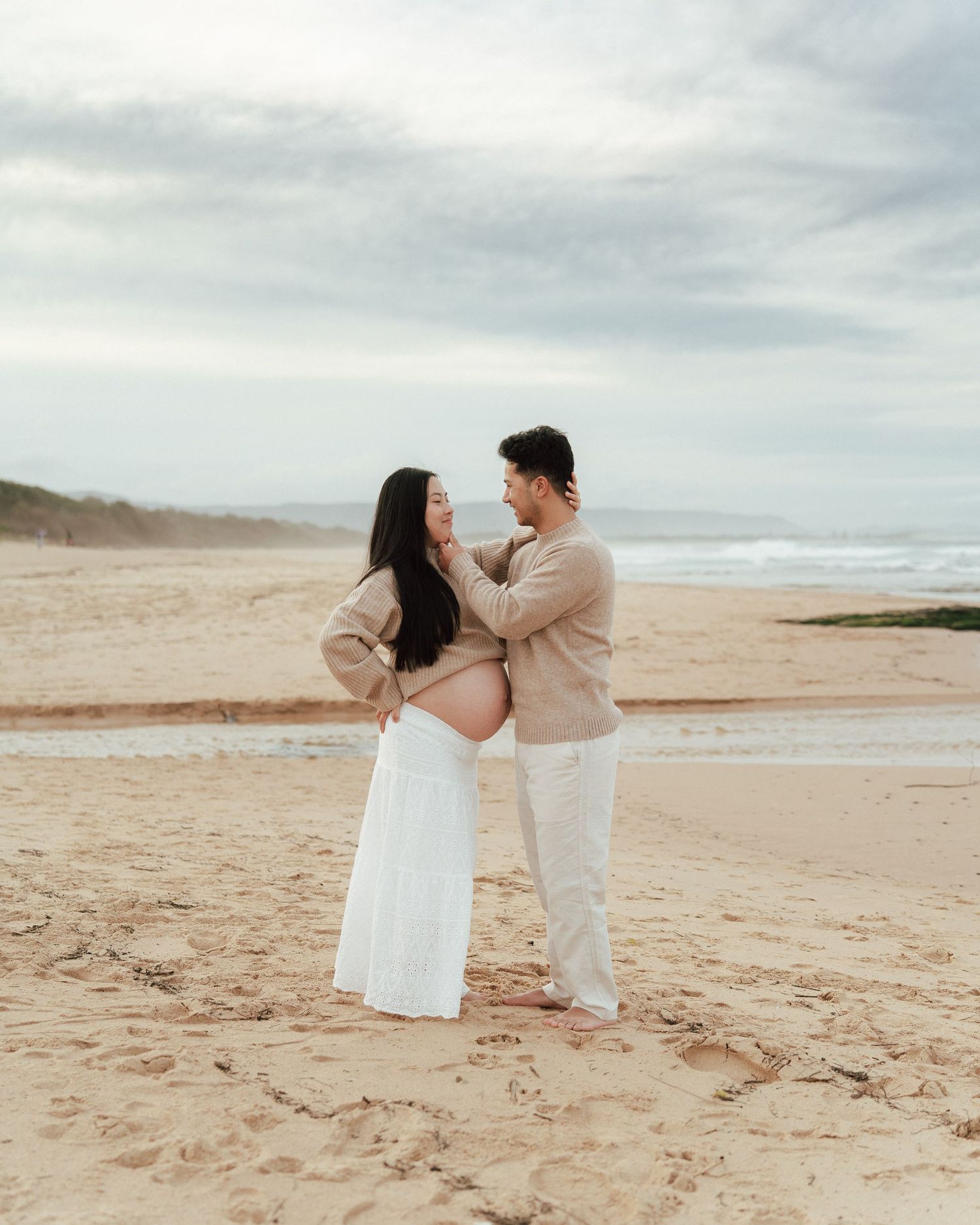 Expecting couple in neutral beige and white tones sharing a tender moment during a beach maternity shoot.