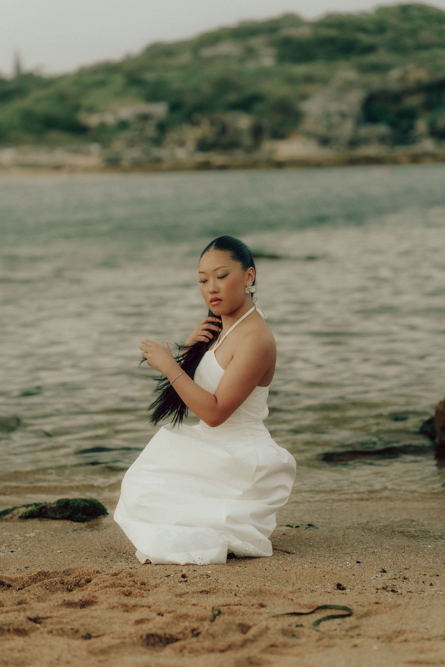 Woman in white halter dress kneeling on sandy beach holding her hair with coastal water behind her.